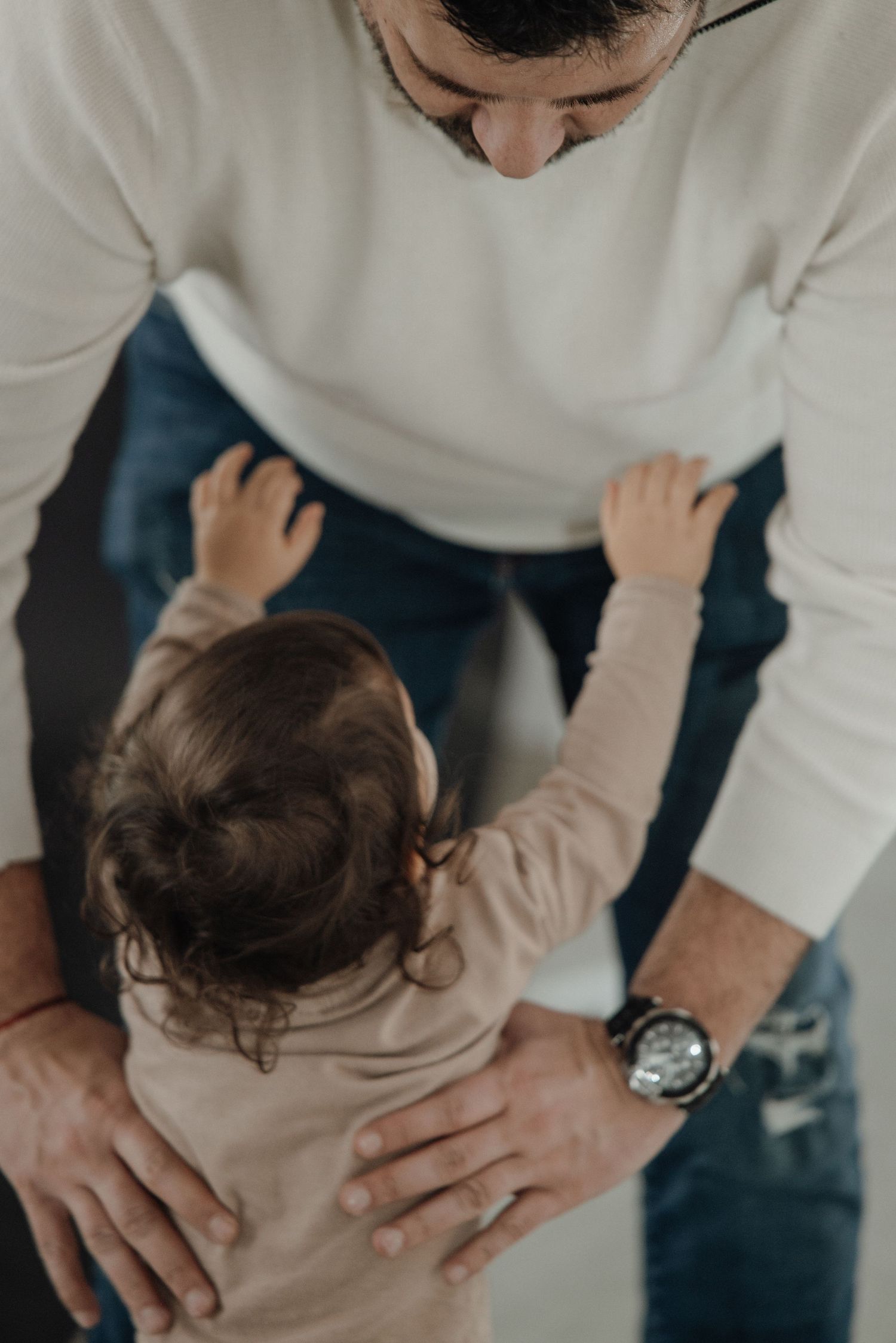 A toddler reaches up to a man in a white sweater and jeans, sharing a tender parent-child moment.