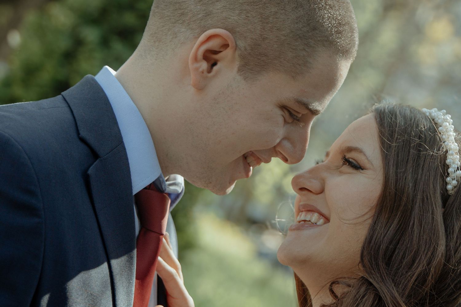 A bride and groom share an intimate moment, nearly touching foreheads, smiling softly outdoors on their wedding day.