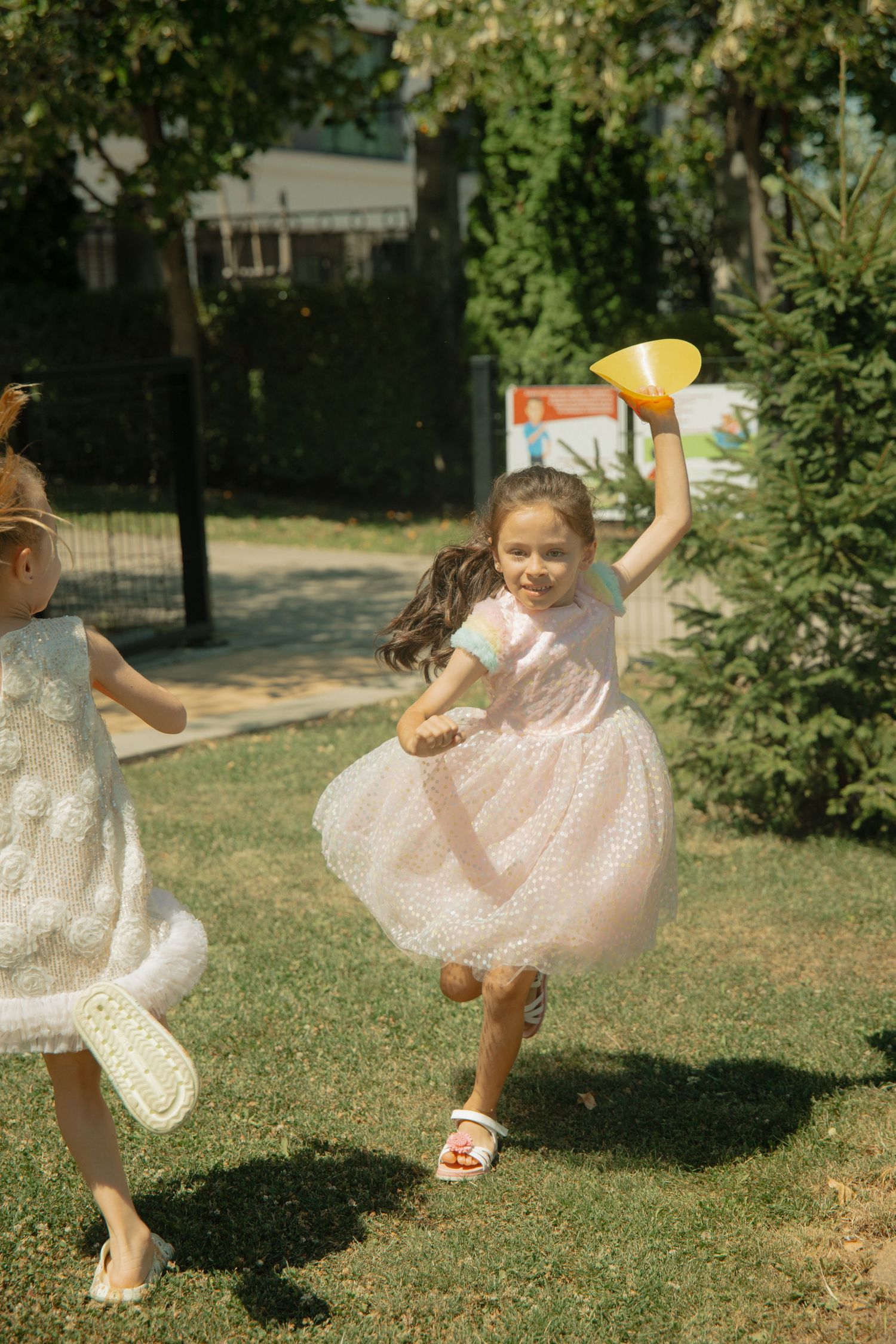 Two girls in party dresses running and playing with a yellow toy in a sunny park.