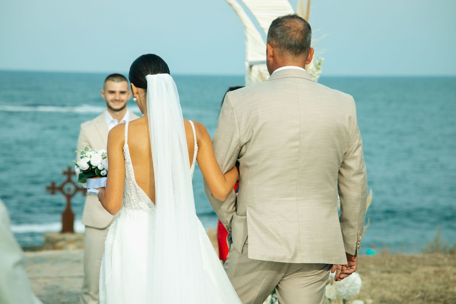 Bride in white gown and veil walks with groom in beige suit toward officiant at outdoor beach wedding ceremony.