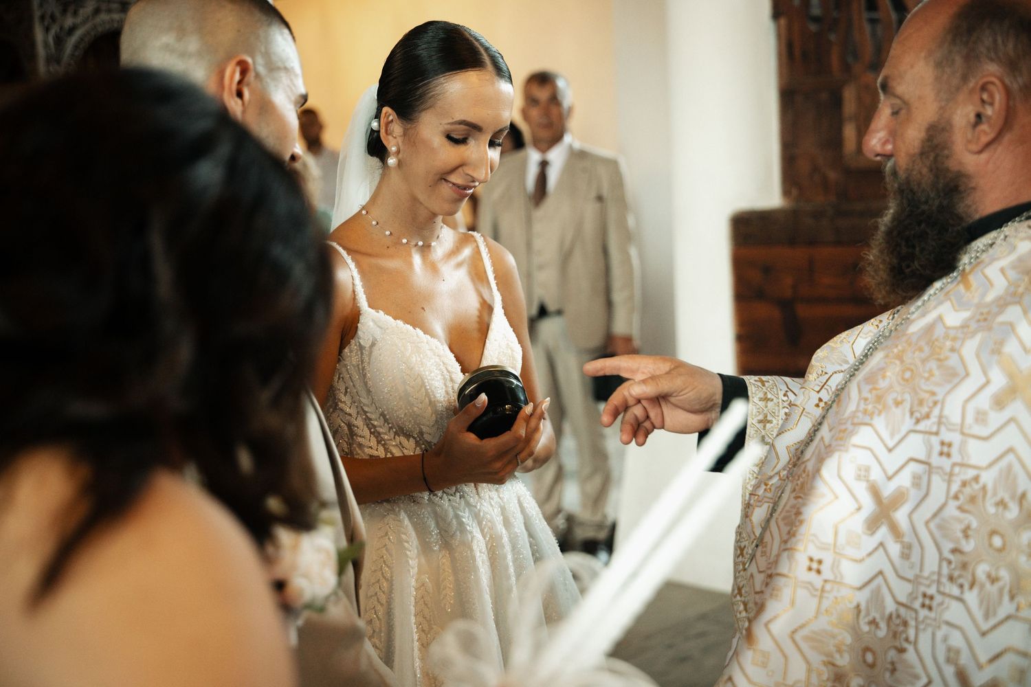 Smiling bride in lace wedding dress receives blessing from Orthodox priest during indoor church wedding ceremony.