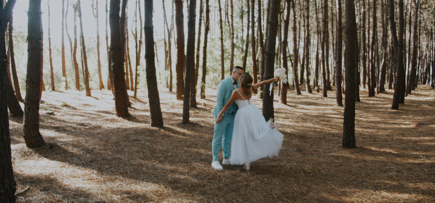 A couple shares a romantic dance in a sunlit pine forest, the bride in white and groom in blue.