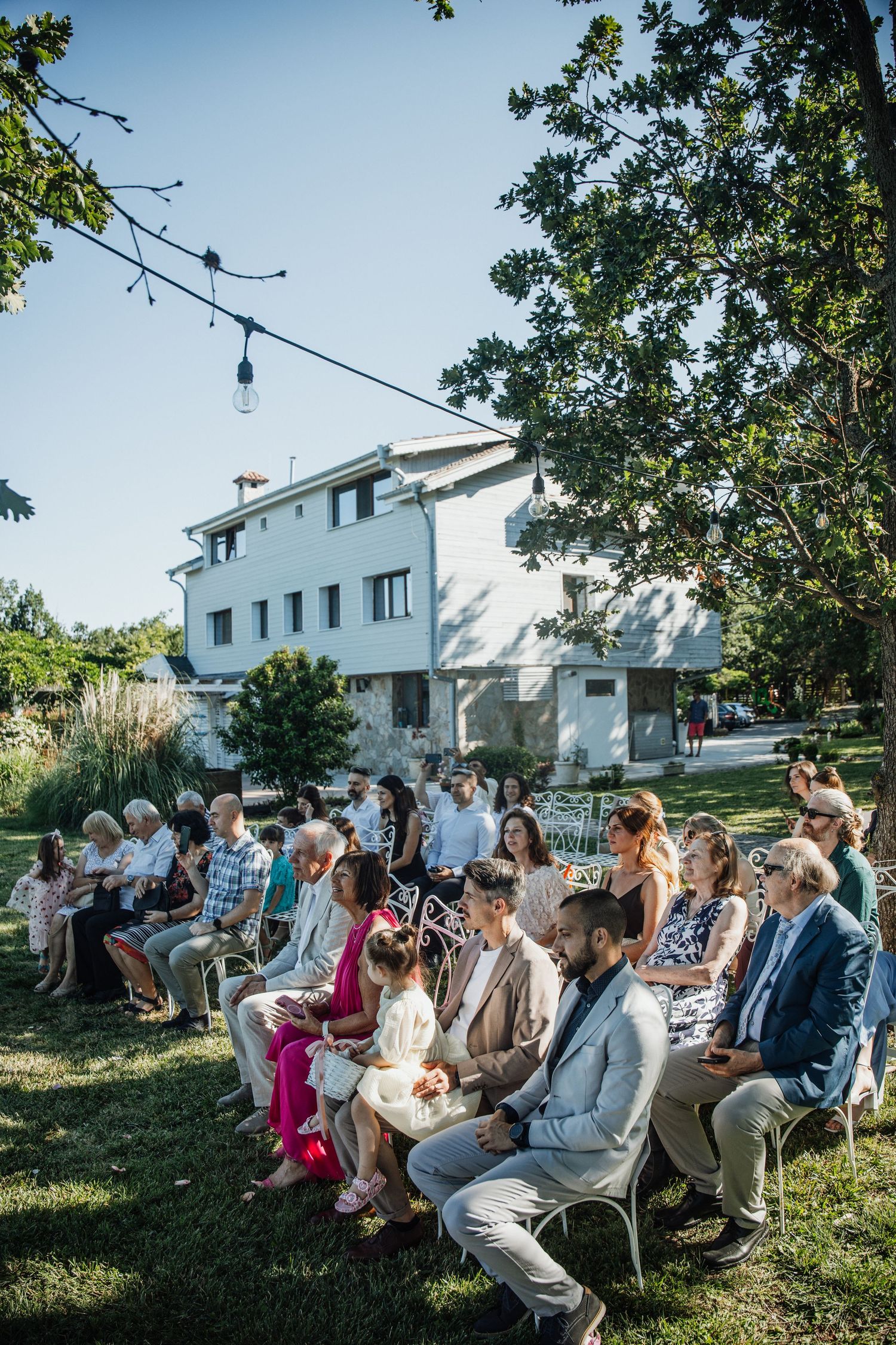Guests seated outdoors on white chairs for a garden wedding ceremony with string lights and white house backdrop.