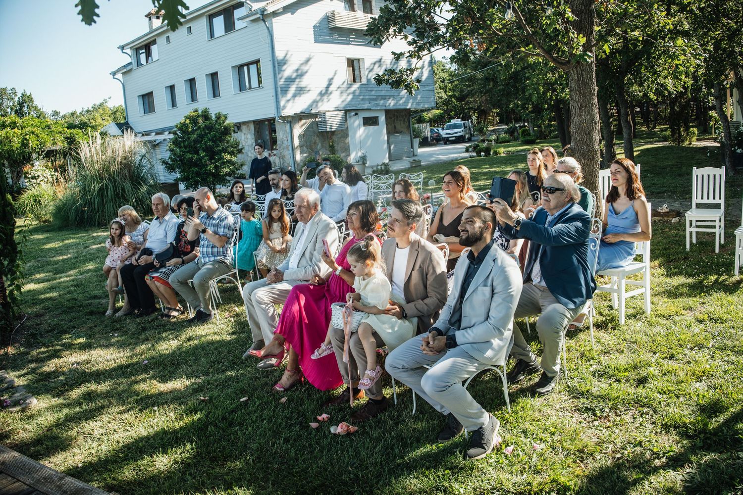 Wedding guests seated on chairs in a sunny garden ceremony with lush trees and a white villa in background.