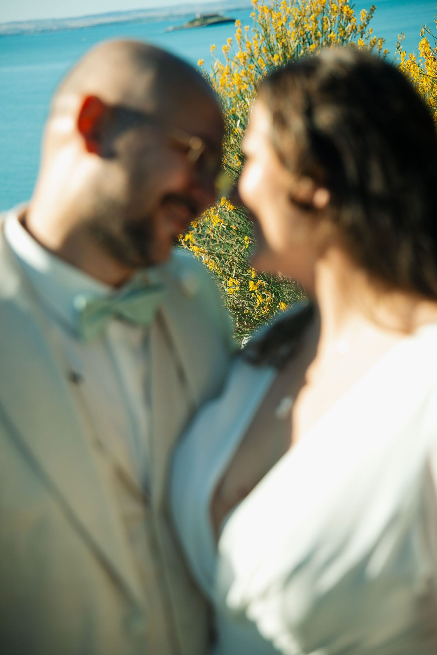 Blurred close-up of a smiling bald groom in a light suit and bride in white near yellow flowers by the sea.