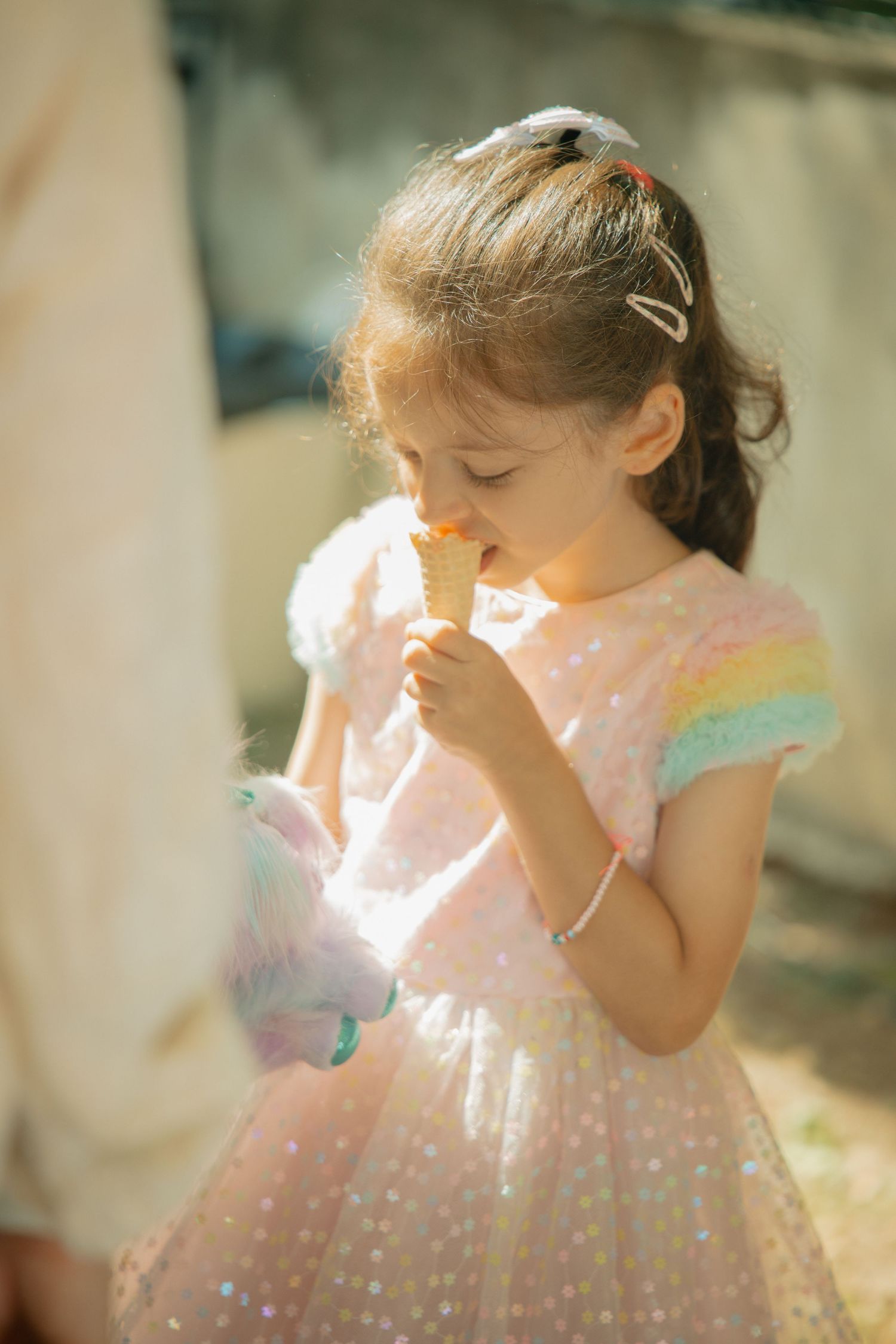 Young girl in pink sequin dress eating ice cream cone outdoors in sunny weather.