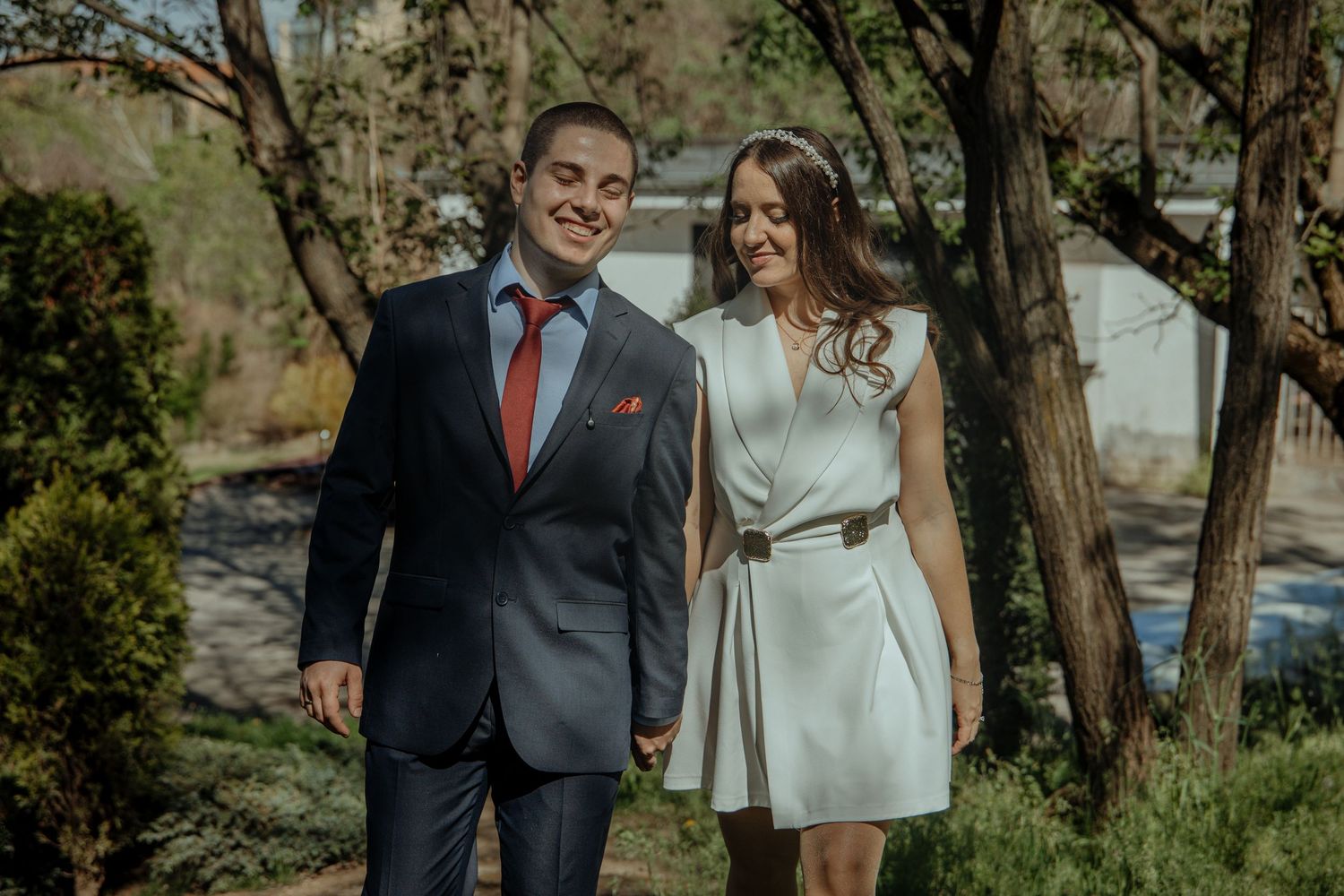 Smiling couple holding hands outdoors, man in navy suit with red tie, woman in white belted dress, surrounded by trees.