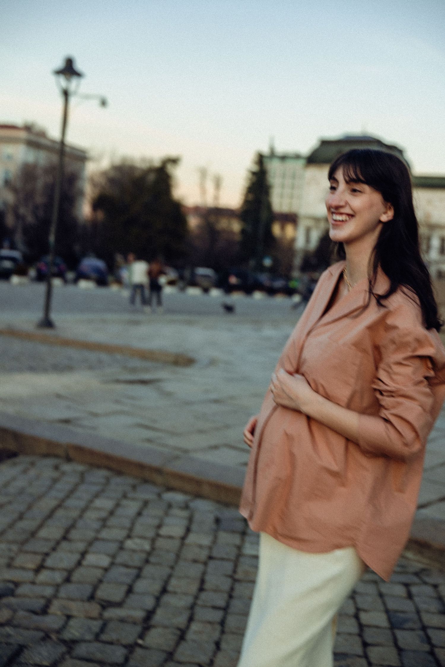 A person in a peach-colored jacket and white skirt poses playfully on a cobblestone street.