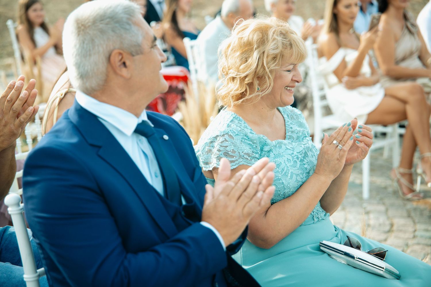 Elegantly dressed wedding guests in blue and teal outfits clapping during outdoor wedding ceremony.