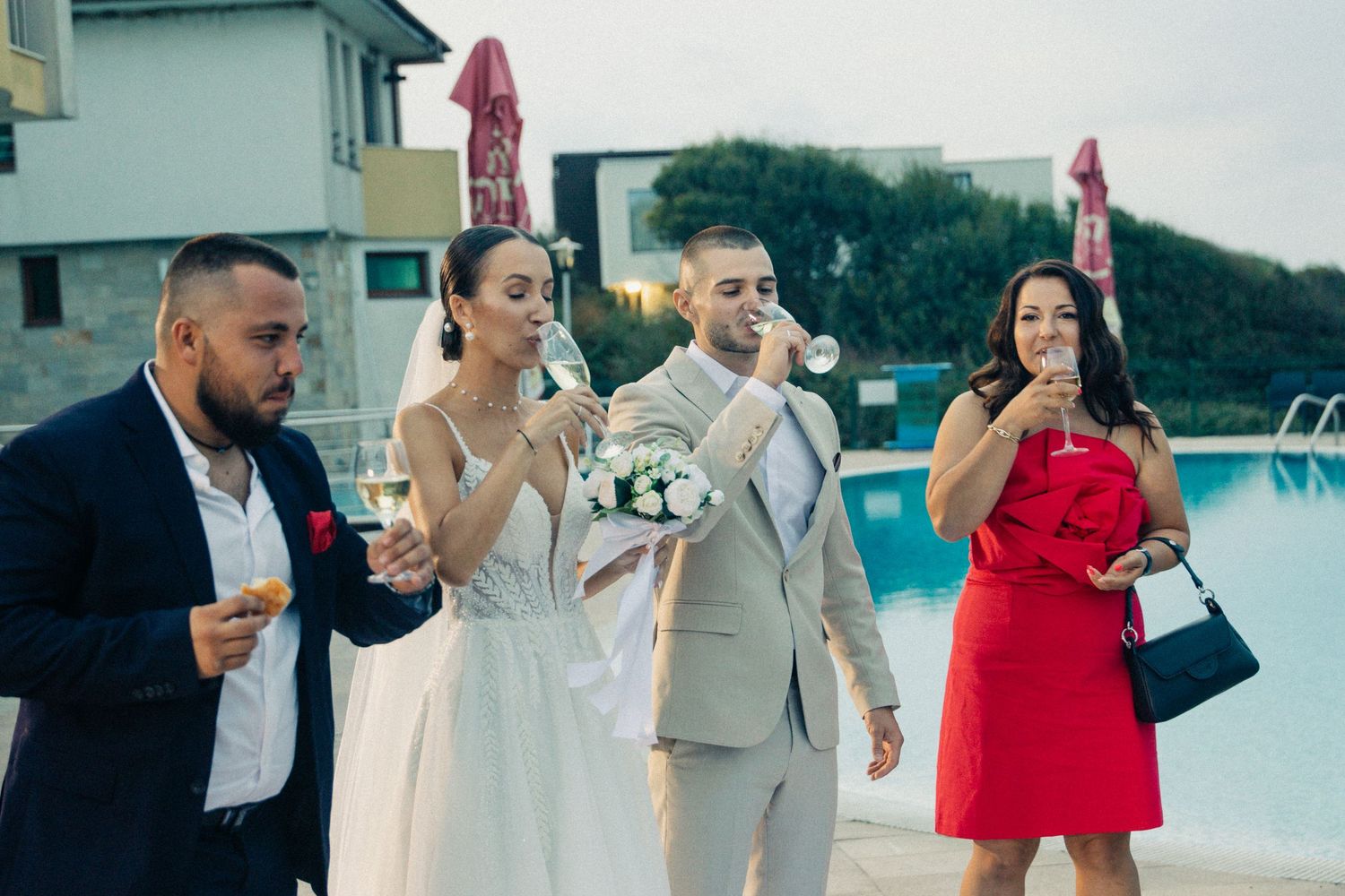 Wedding couple with guests celebrating outdoors near a pool, bride holding bouquet while guests enjoy drinks.