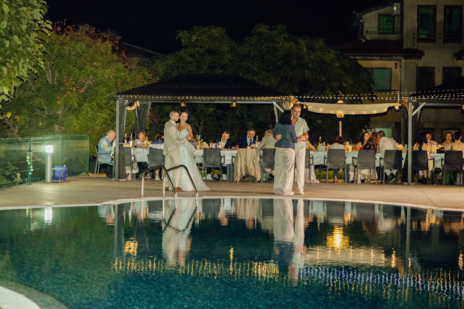Couple shares first dance beside illuminated pool at elegant outdoor wedding reception at night.