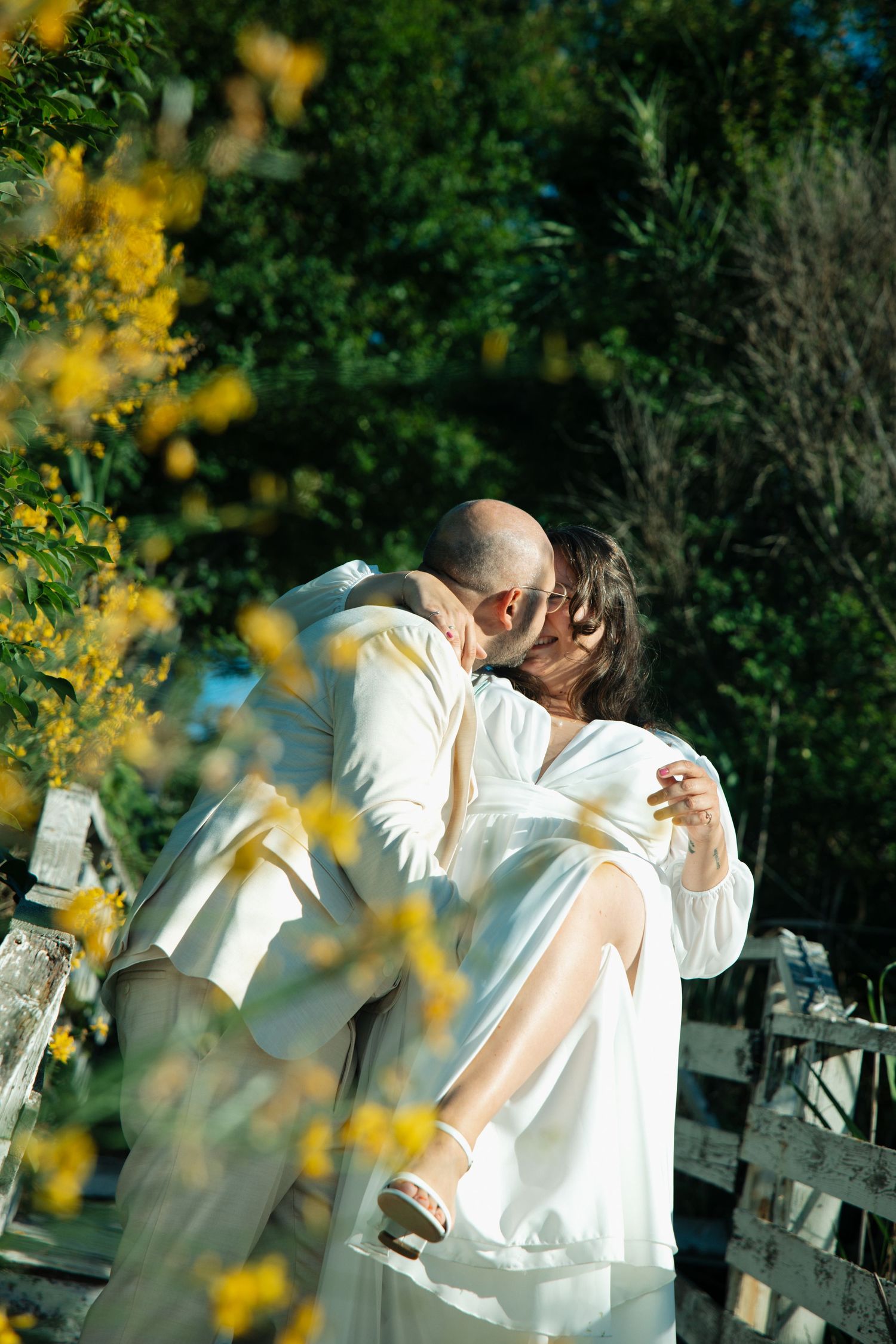 Couple sharing a romantic kiss outdoors, surrounded by yellow flowers and lush green trees in natural light.