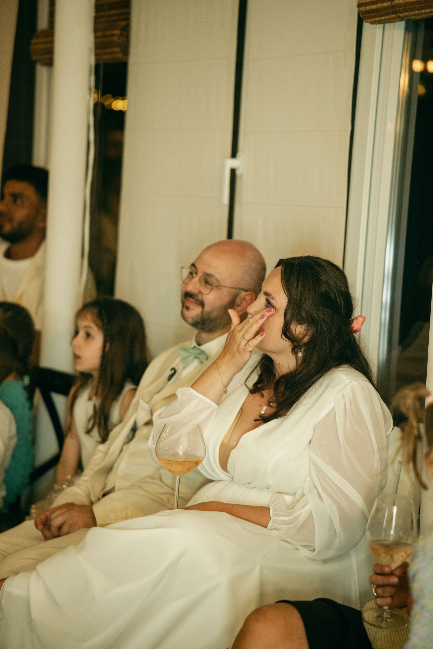 A couple in wedding attire sit together laughing during a reception speech indoors.