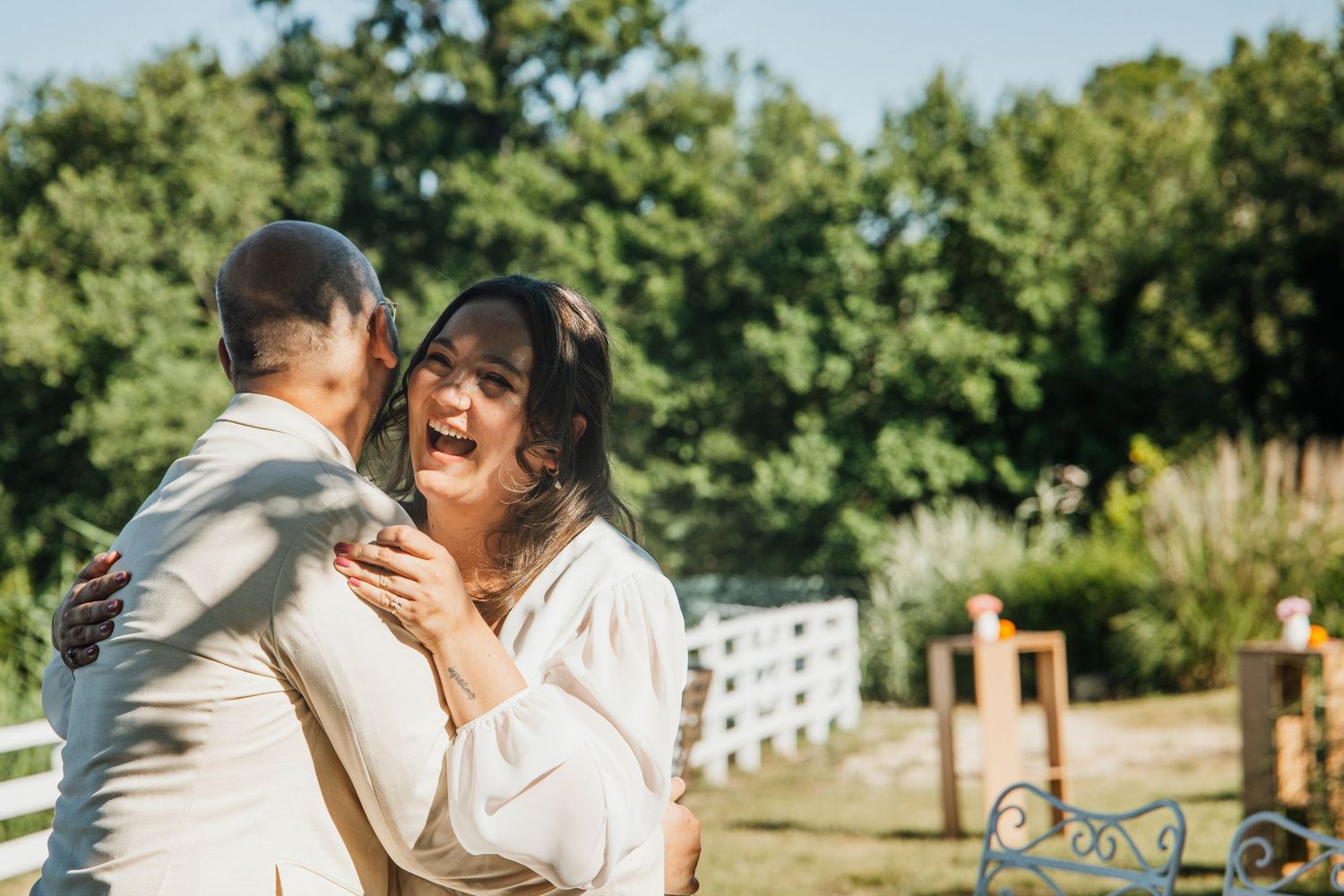 Emotional bride embracing a guest in a white outfit at an outdoor wedding venue with a white fence.