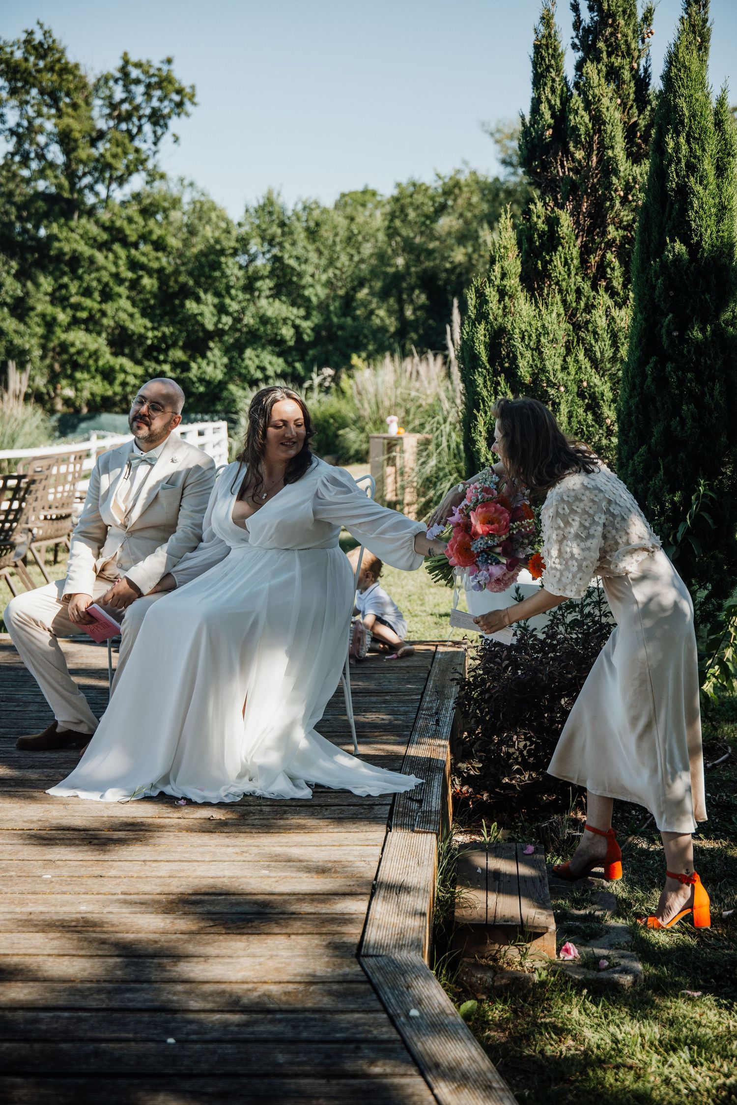 Bride in white gown seated outdoors at garden wedding ceremony with guests on wooden deck.