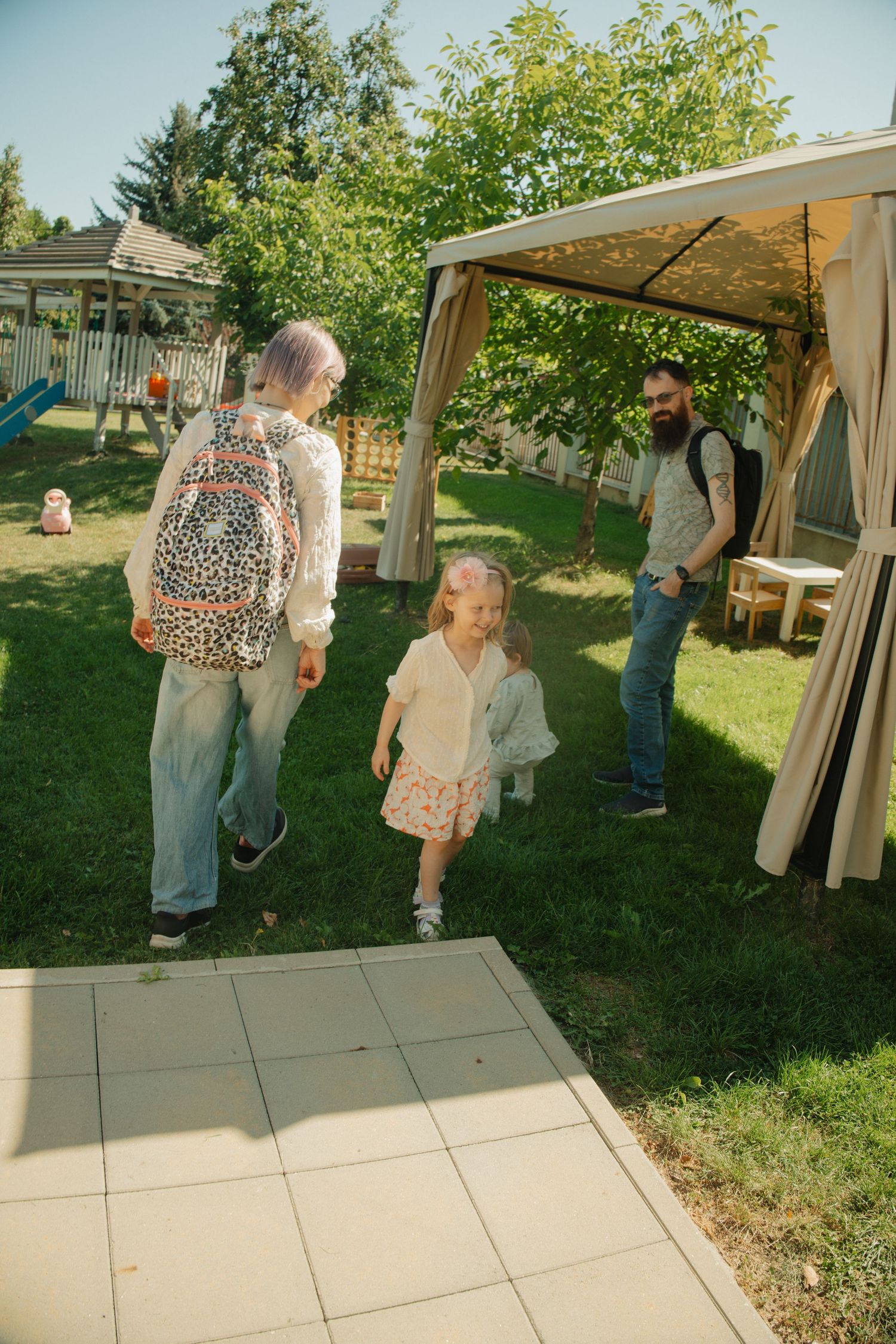 Family with young girl playing in sunny backyard garden near gazebo on a beautiful summer day.