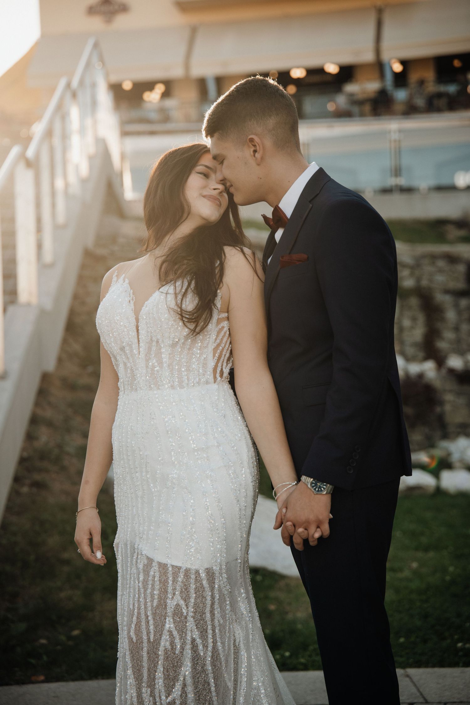 Bride in sequined white gown and groom in black suit hold hands and touch foreheads at golden hour outdoor wedding.