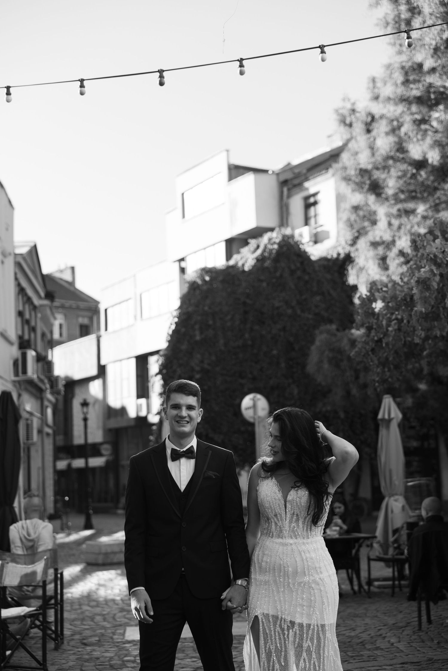 Black and white photo of a stylish wedding couple holding hands on a cobblestone street with string lights overhead.