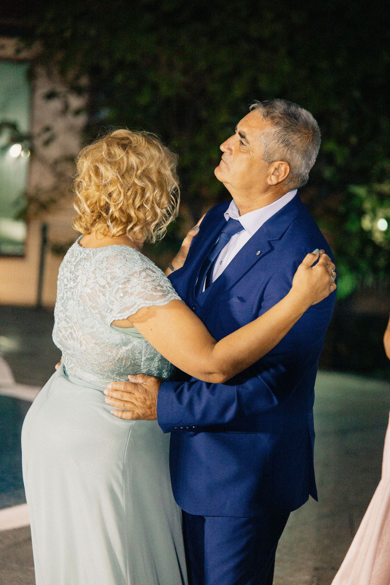 Older couple dancing together outdoors at night, man in blue suit, woman in sage green lace dress.