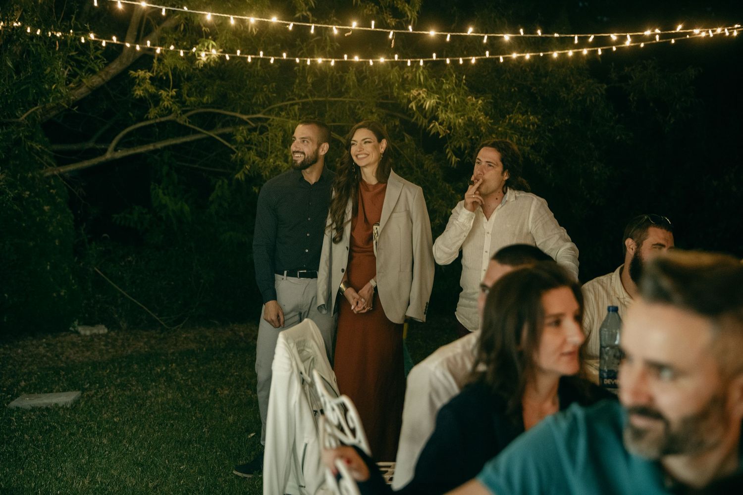 Wedding guests laugh and socialize under string lights at an outdoor evening reception.