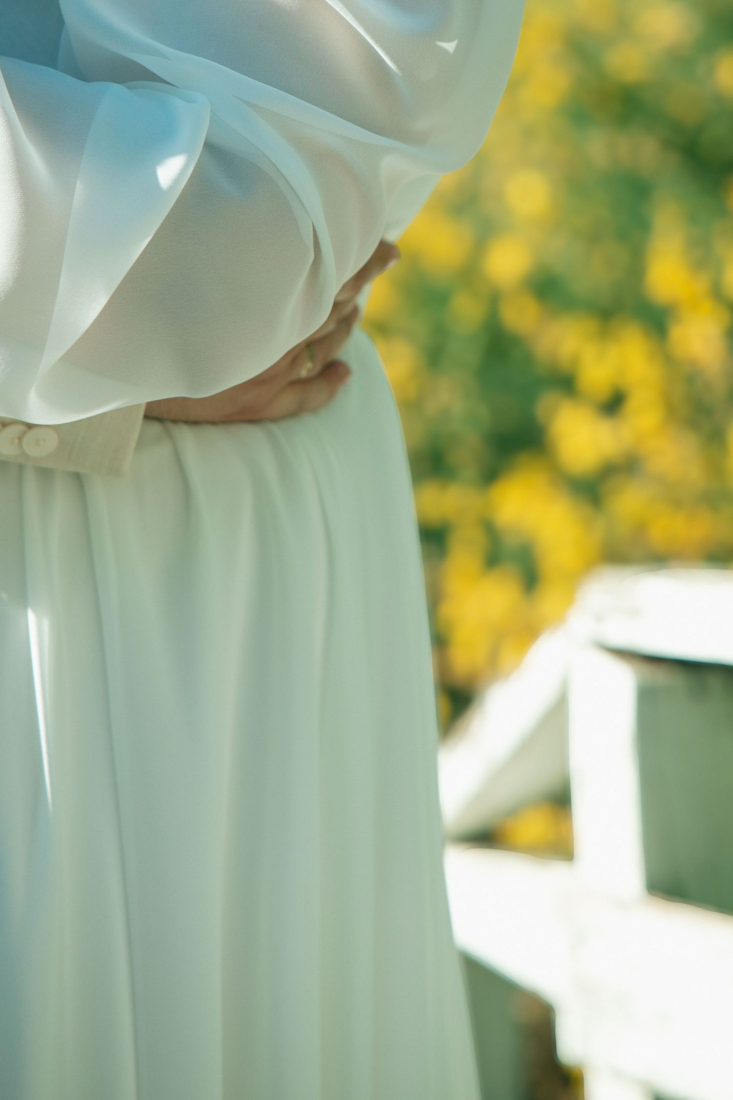 Pregnant woman in white dress cradling baby bump outdoors with yellow flowers in background.