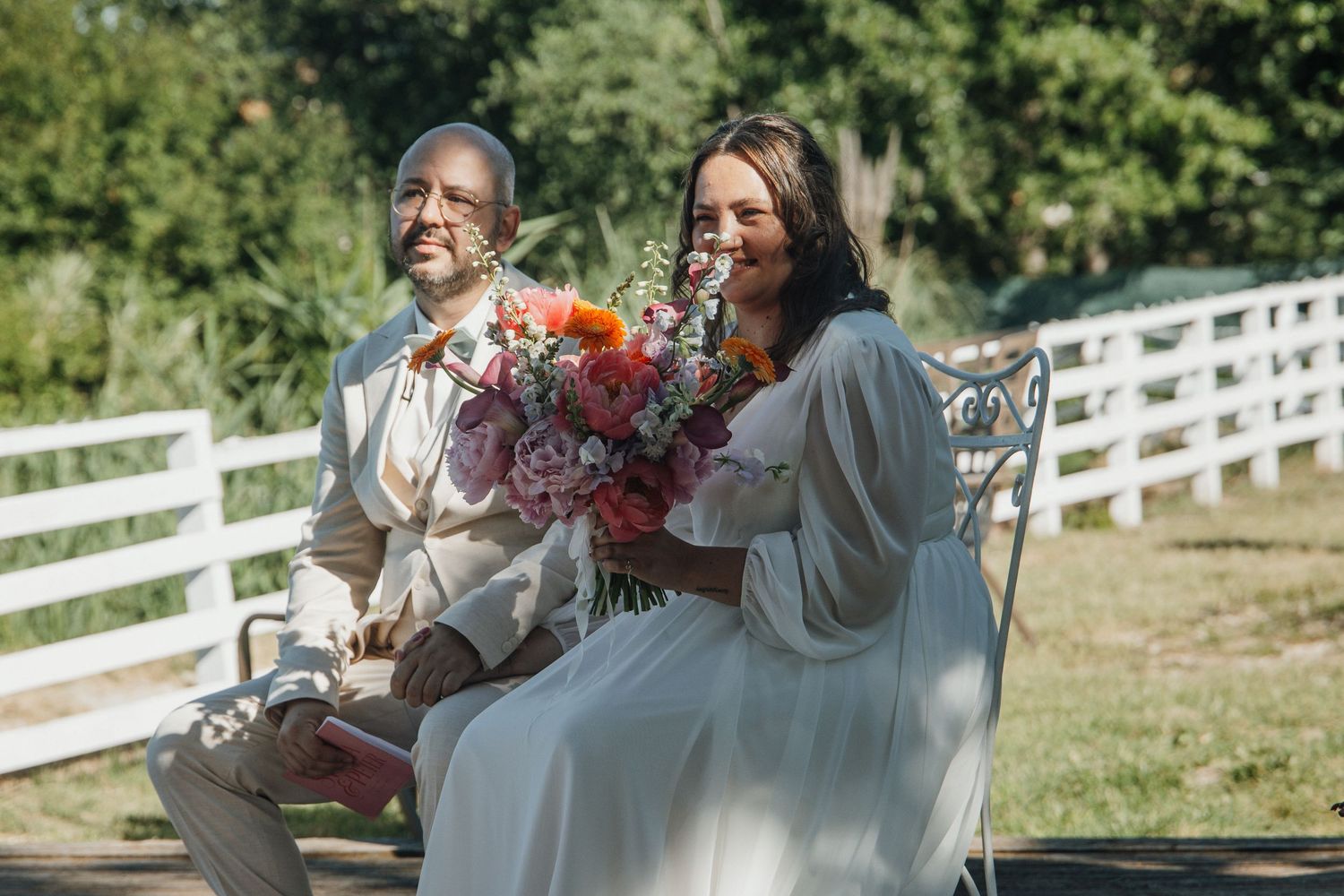 Bride in white gown sits with elderly man on bench holding vibrant floral bouquet outdoors.