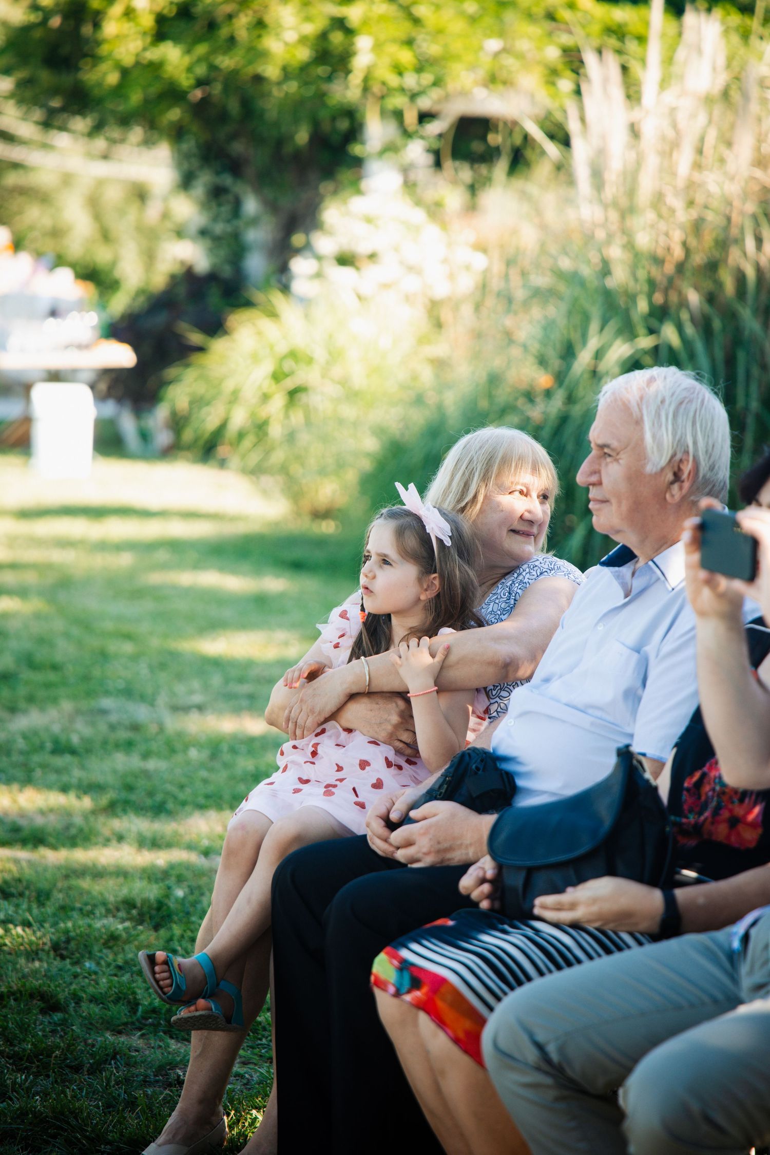 Grandparents sit with young girl on bench watching outdoor wedding ceremony in garden.
