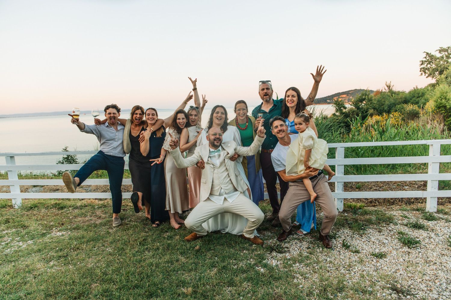 Group of friends posing and celebrating outdoors near a white fence with ocean views at sunset.