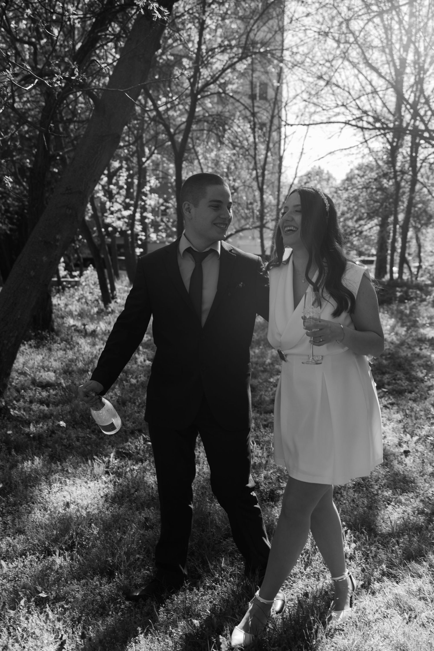 Black and white photo of smiling bride and groom walking through a sunlit park on wedding day.