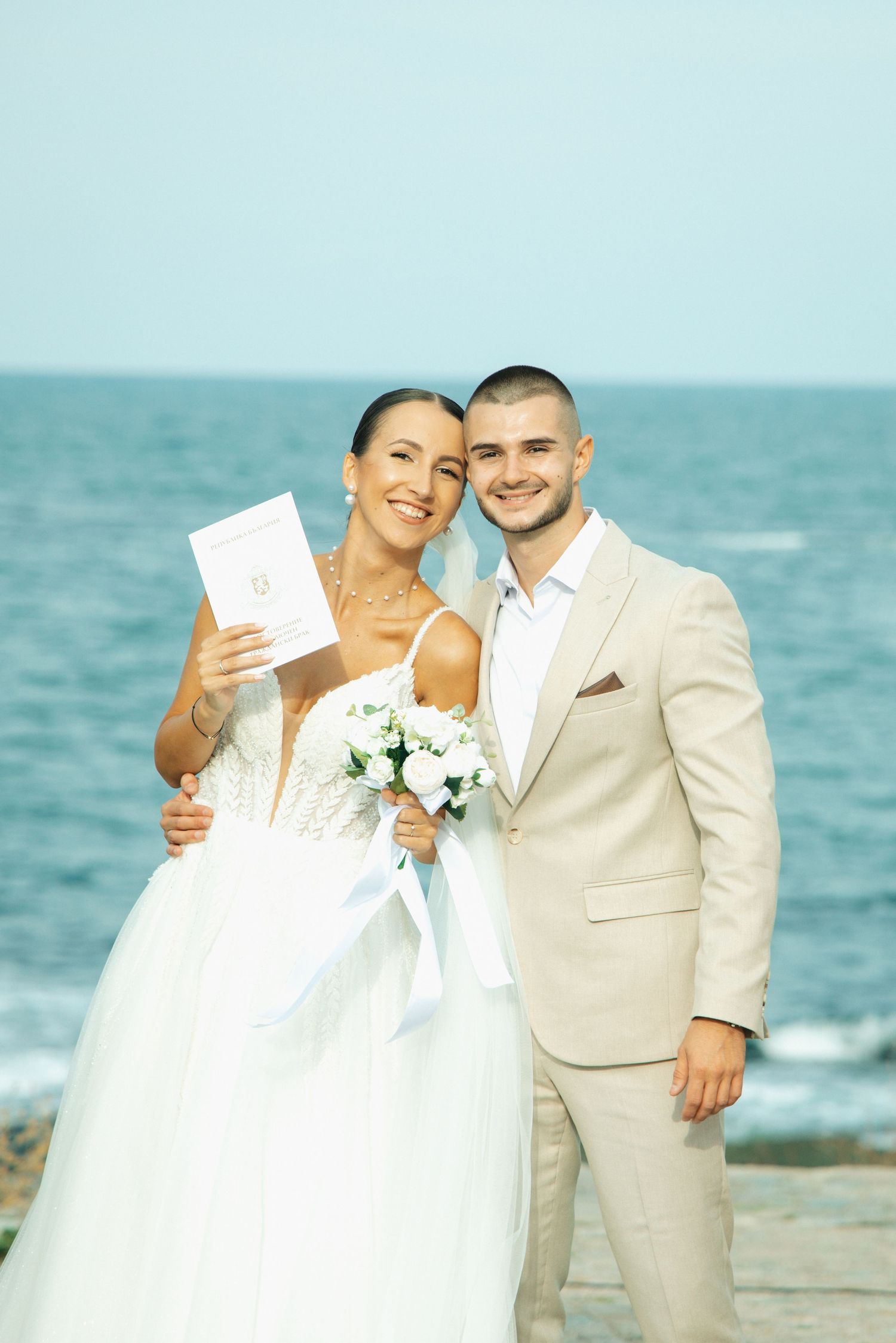 Happy newlyweds pose with marriage certificate and white bouquet with ocean backdrop on wedding day.