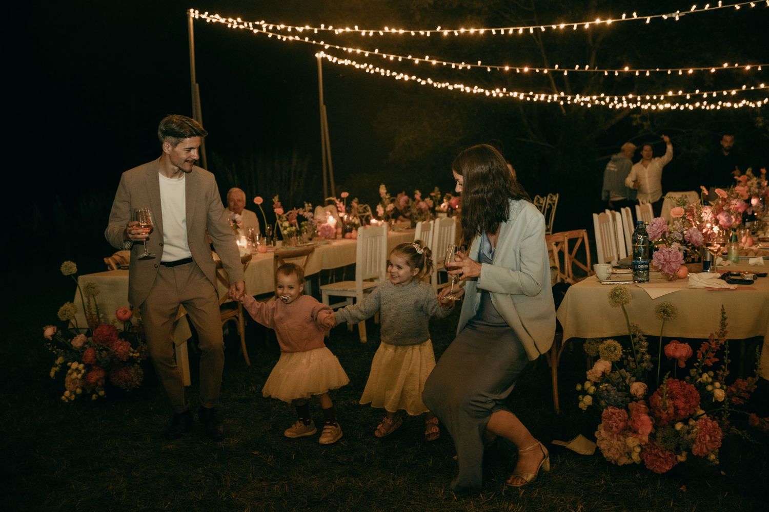Family dances joyfully at night under string lights at an outdoor wedding reception with floral arrangements.