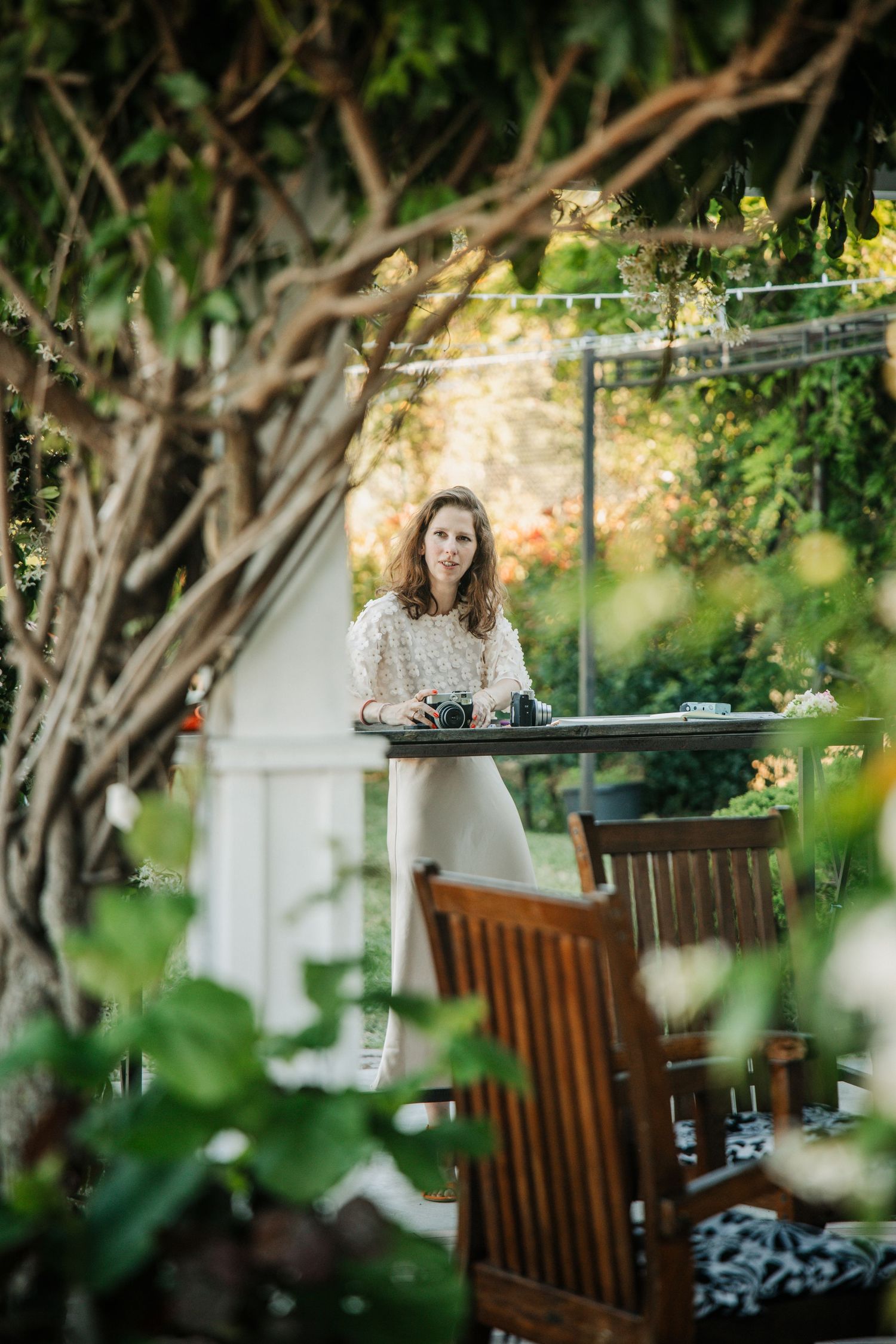 A curly-haired woman in a white lace dress stands at an outdoor table surrounded by lush green garden foliage.
