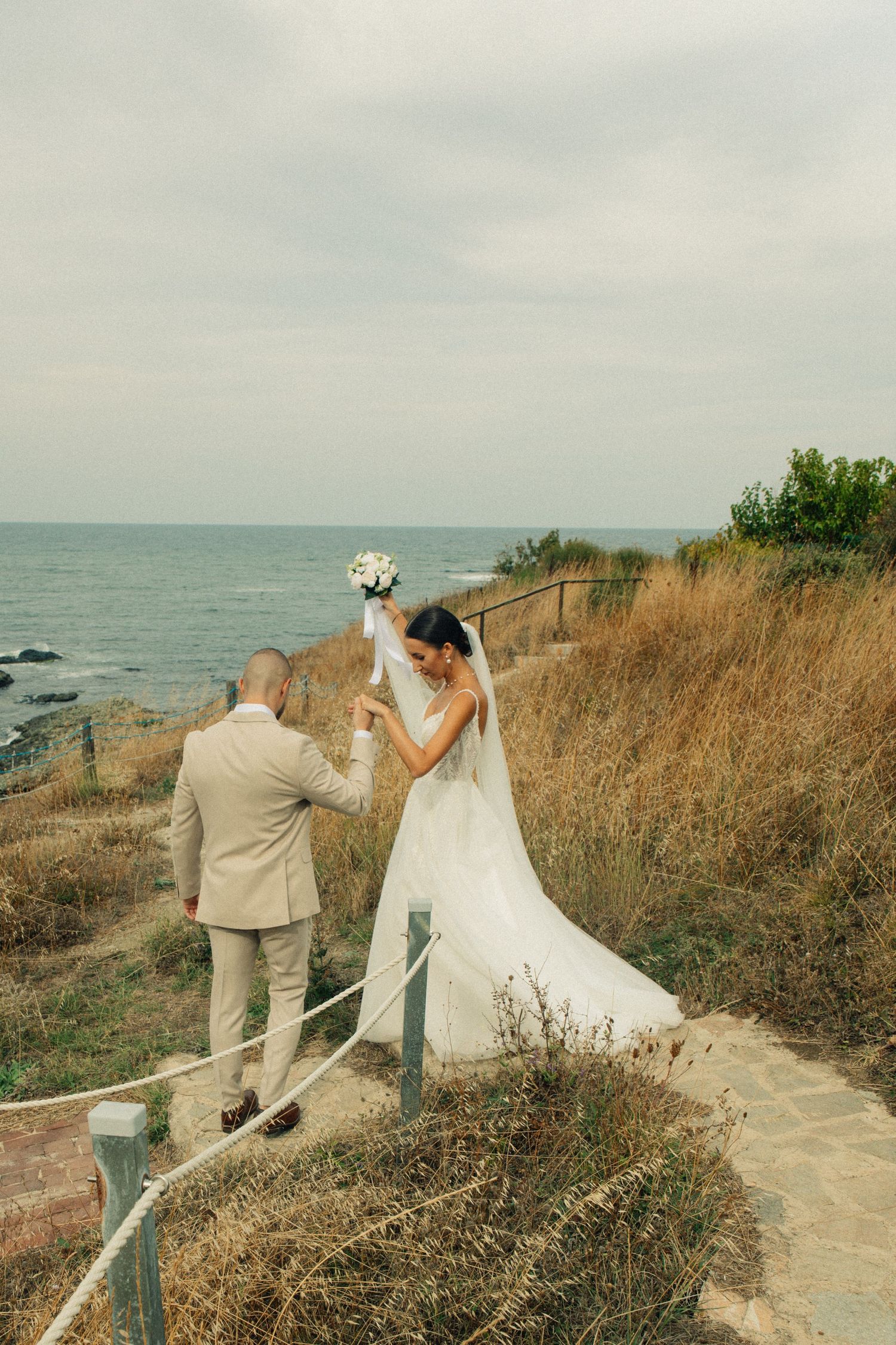 Bride and groom walk along a coastal path, bride in white gown holding bouquet, groom in beige suit, ocean backdrop.