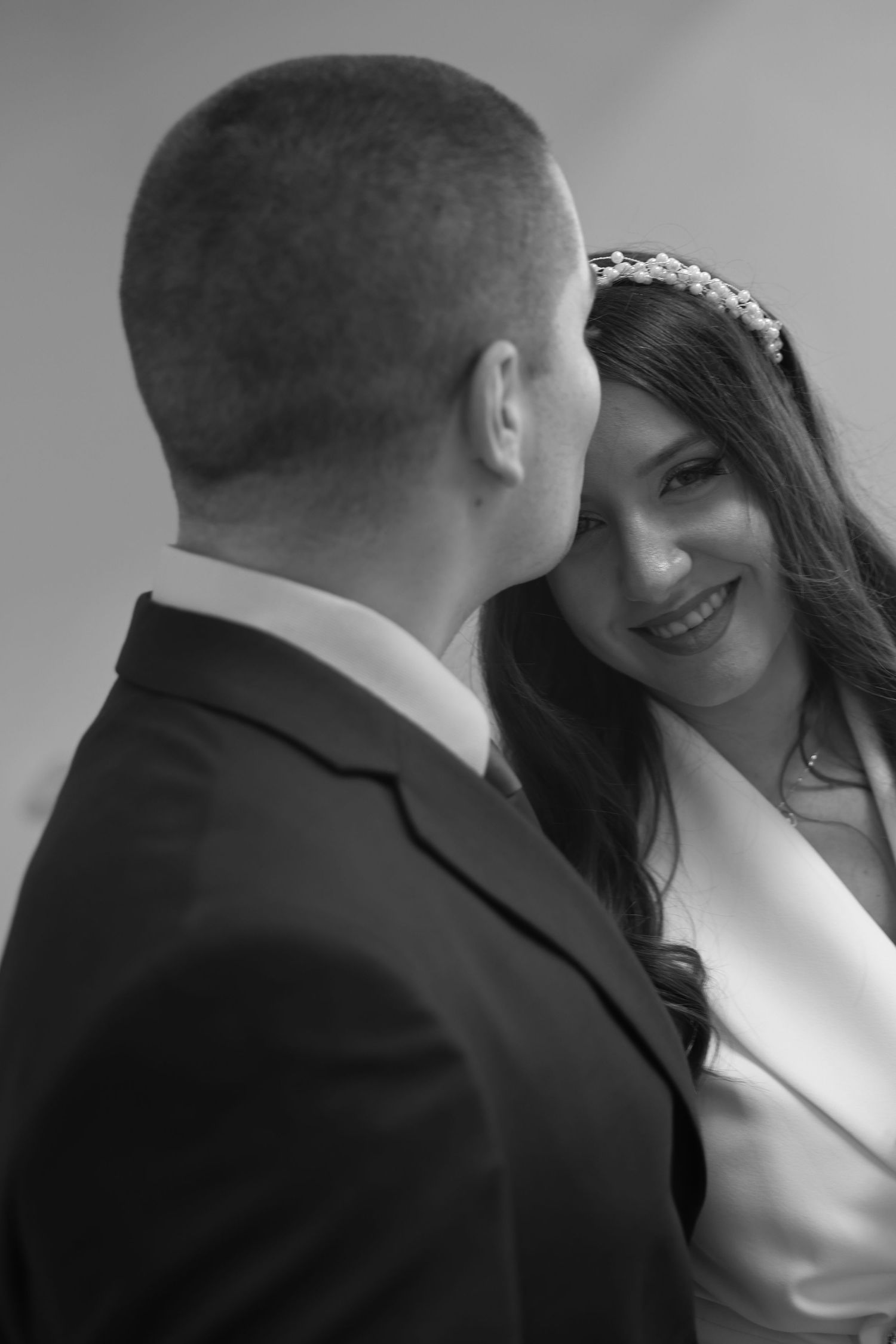Black and white wedding photo of smiling bride in tiara gazing at groom in dark suit.