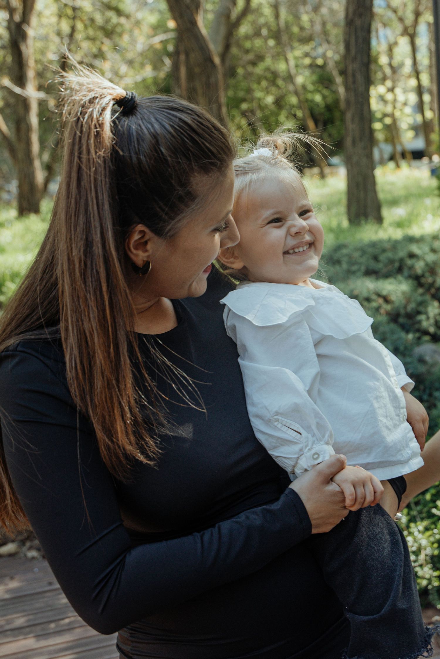 Young mother in black top holds laughing toddler girl in white ruffled blouse outdoors in a sunlit park.