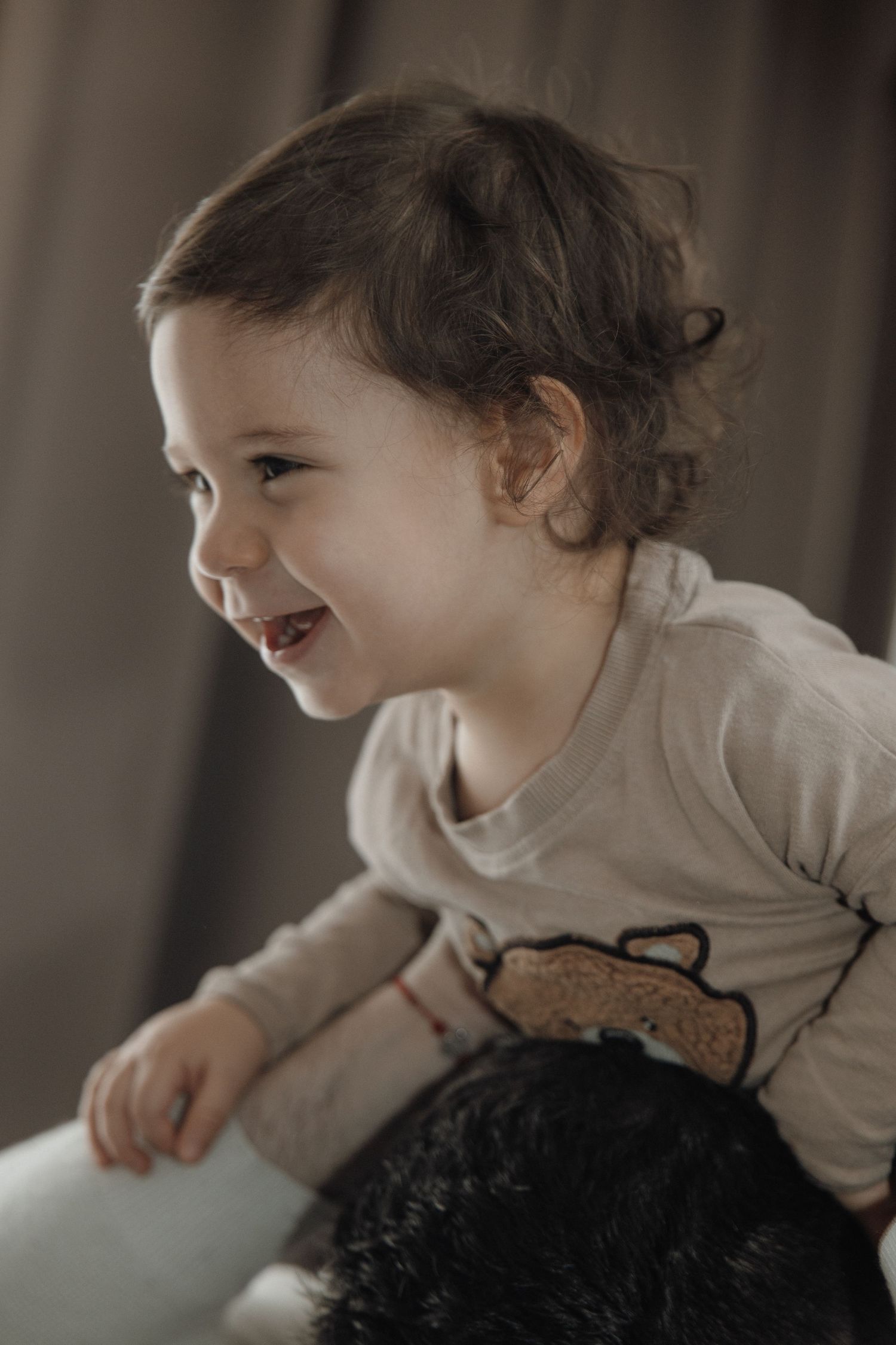 A joyful toddler with curly hair laughs while wearing a bear-print shirt, leaning forward with delight.