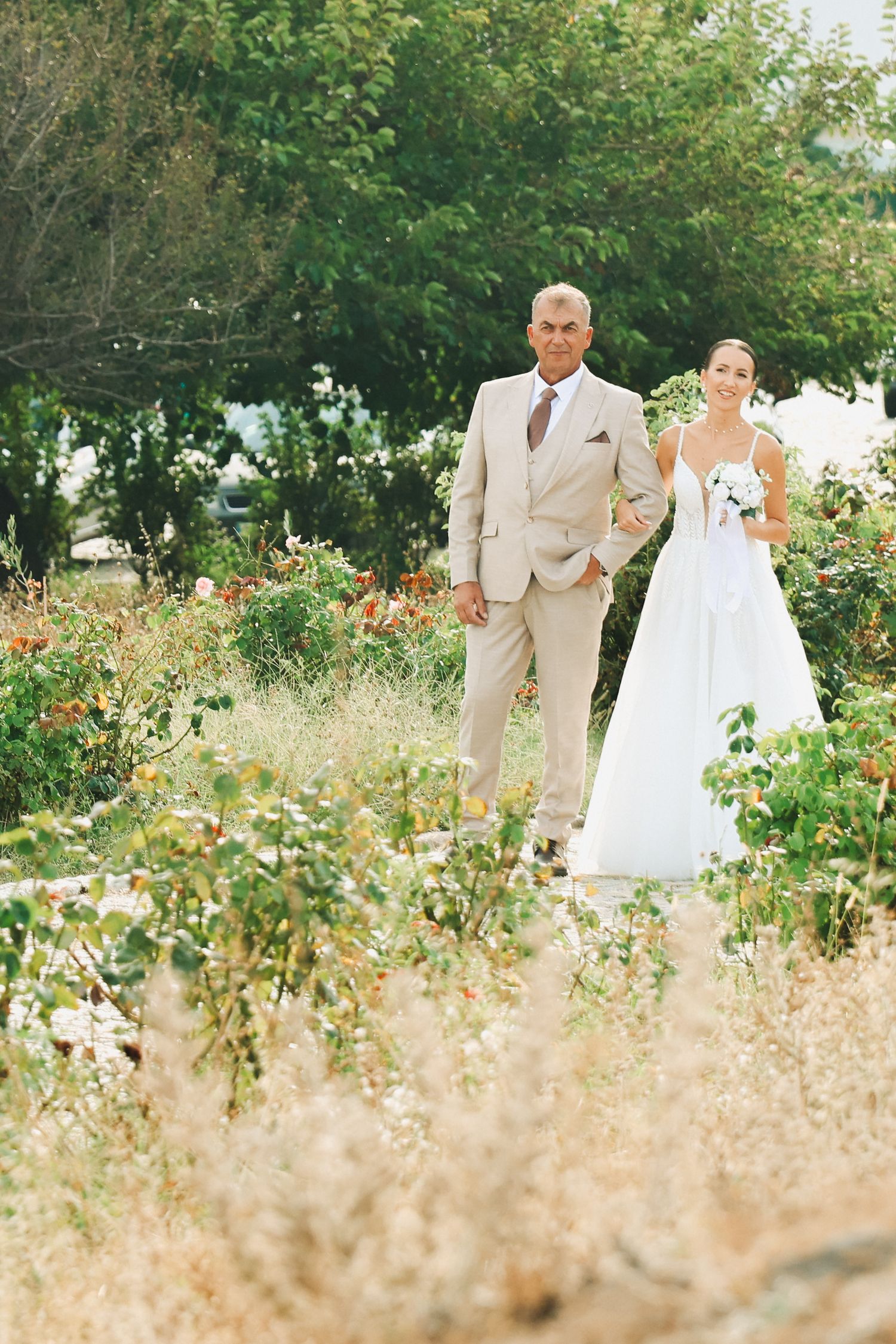 Bride and groom walking through a lush garden, bride in white gown holding bouquet, groom in beige suit.