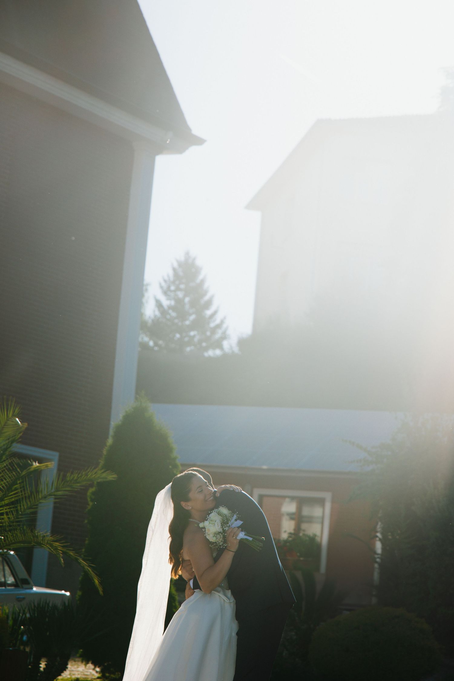 Bride and groom share a romantic kiss outdoors in soft sunlight, with lush greenery and elegant buildings in the background.