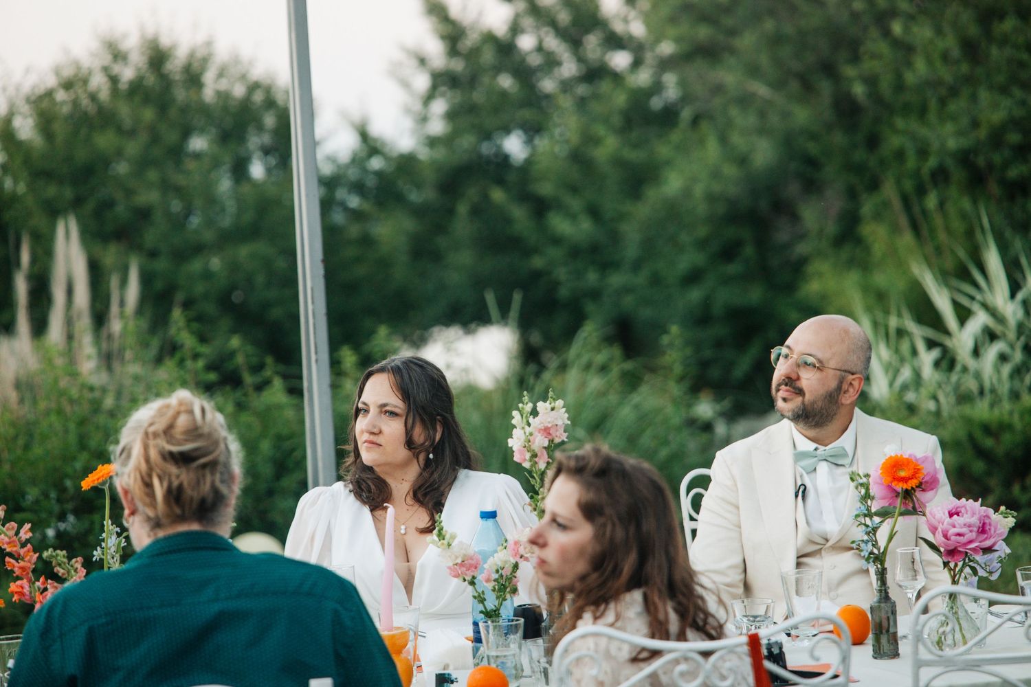 Bride and groom with glasses sit at decorated outdoor wedding reception table surrounded by colorful flowers.