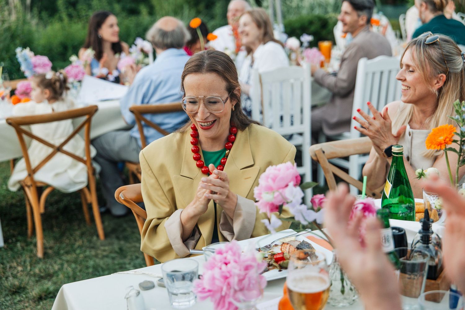 Woman in yellow blazer laughing at outdoor garden party with floral decorations and guests dining.