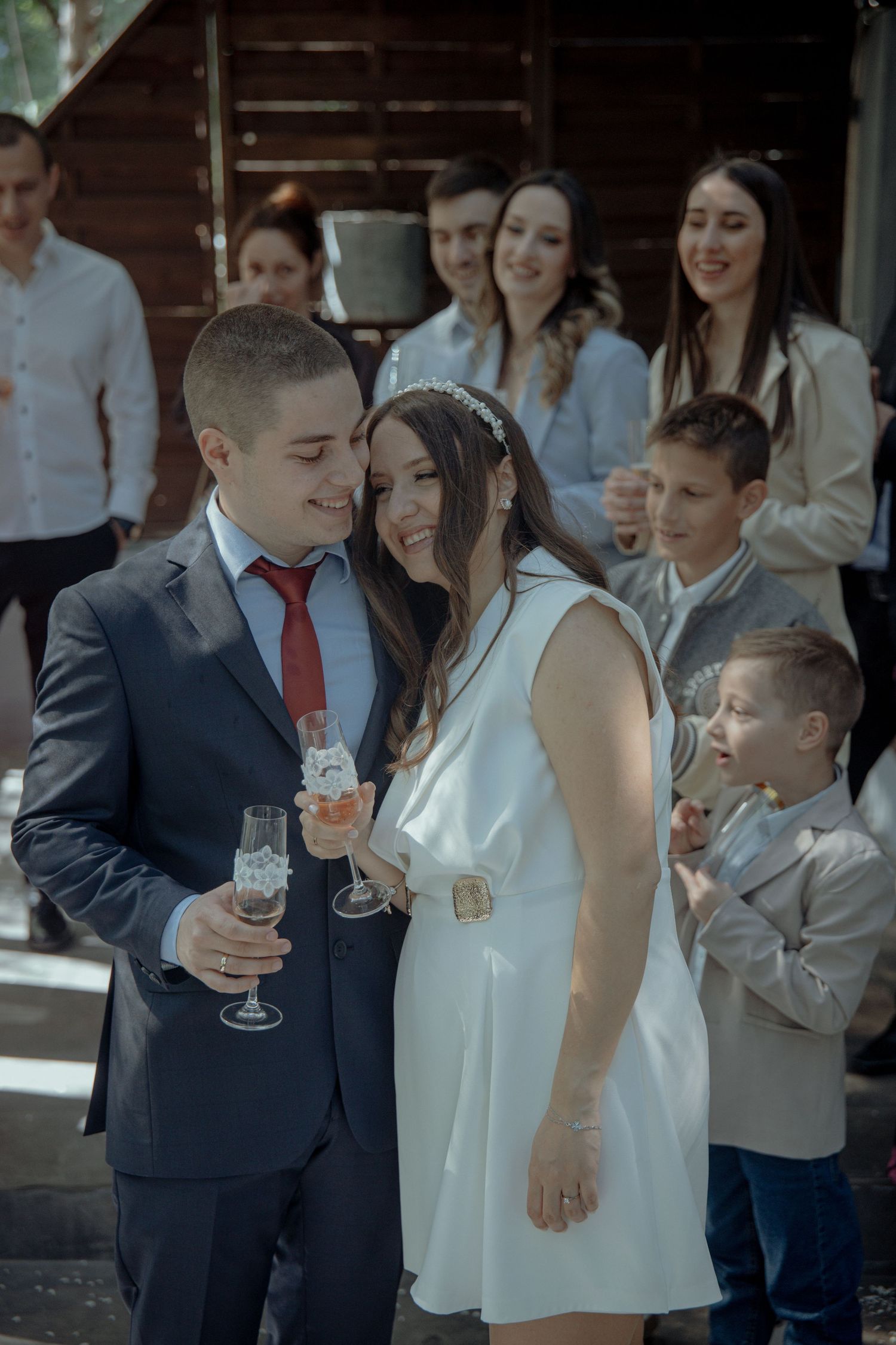 Newlywed couple holding champagne glasses smiling at wedding reception with guests in background.