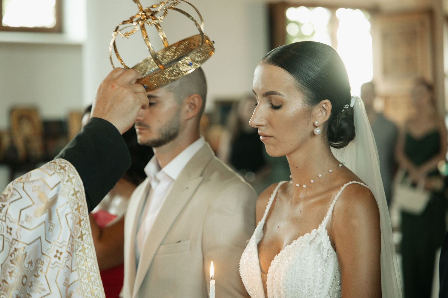 Orthodox priest placing golden crown on groom during traditional wedding ceremony with candlelit bride in veil.