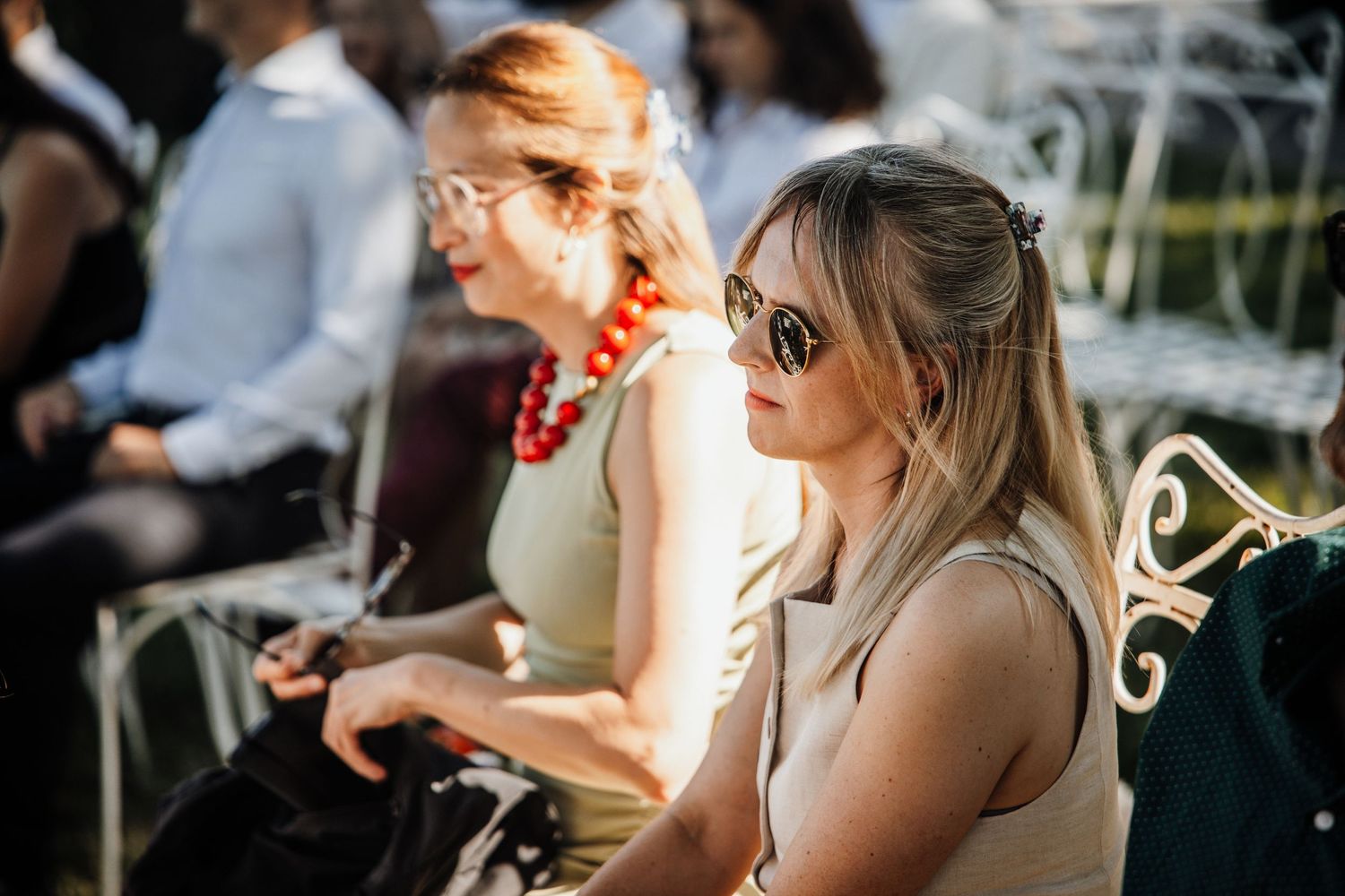 Two elegantly dressed women seated at outdoor wedding, one wearing red beaded necklace, other in sunglasses.
