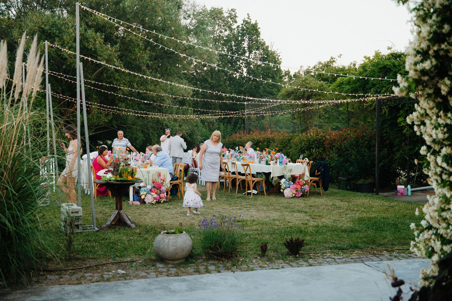 Outdoor garden wedding reception with string lights overhead and guests gathered around long decorated tables at dusk.