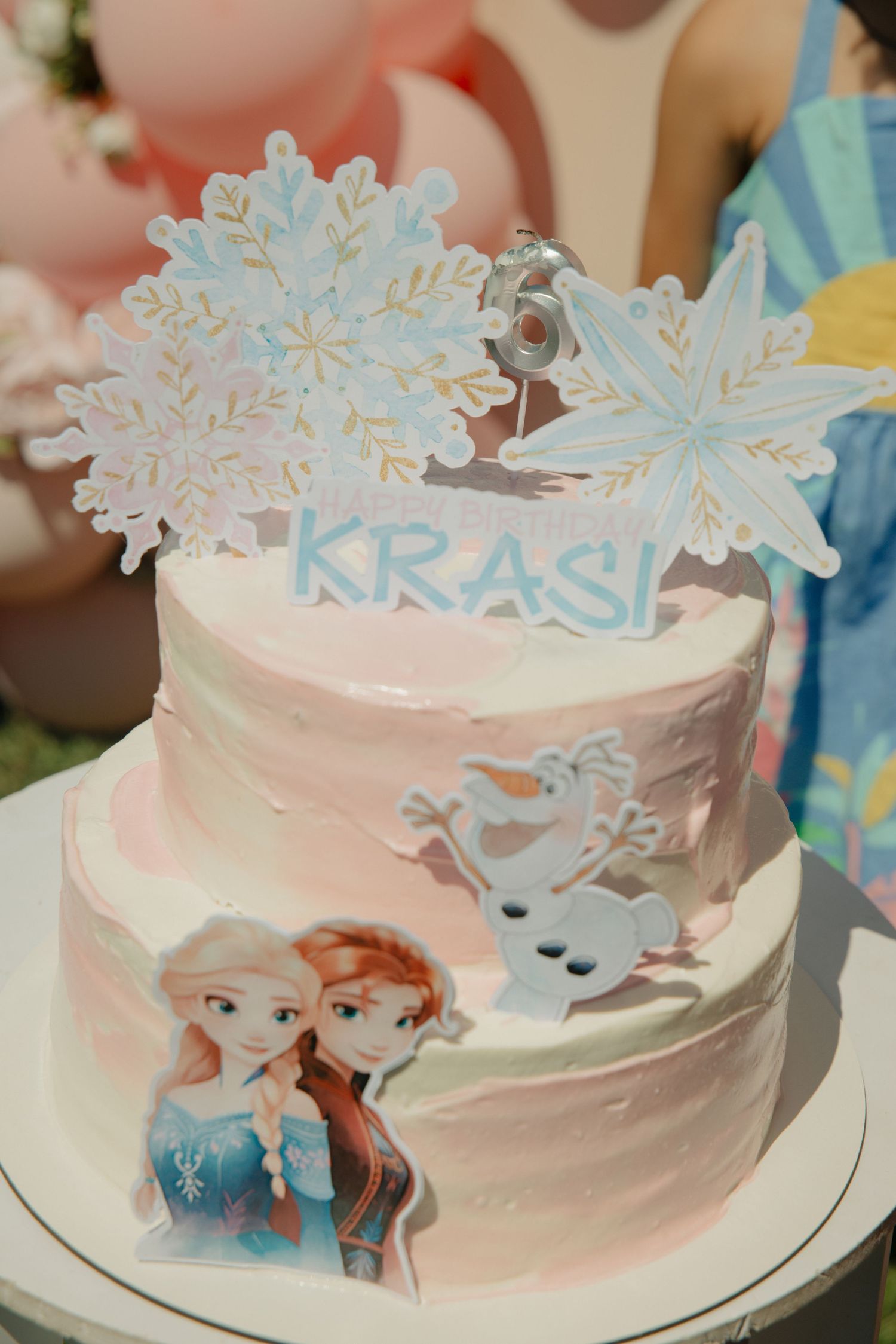 Frozen-themed two-tier pink birthday cake with Elsa, Anna, Olaf decorations and snowflake topper reading KRASI.