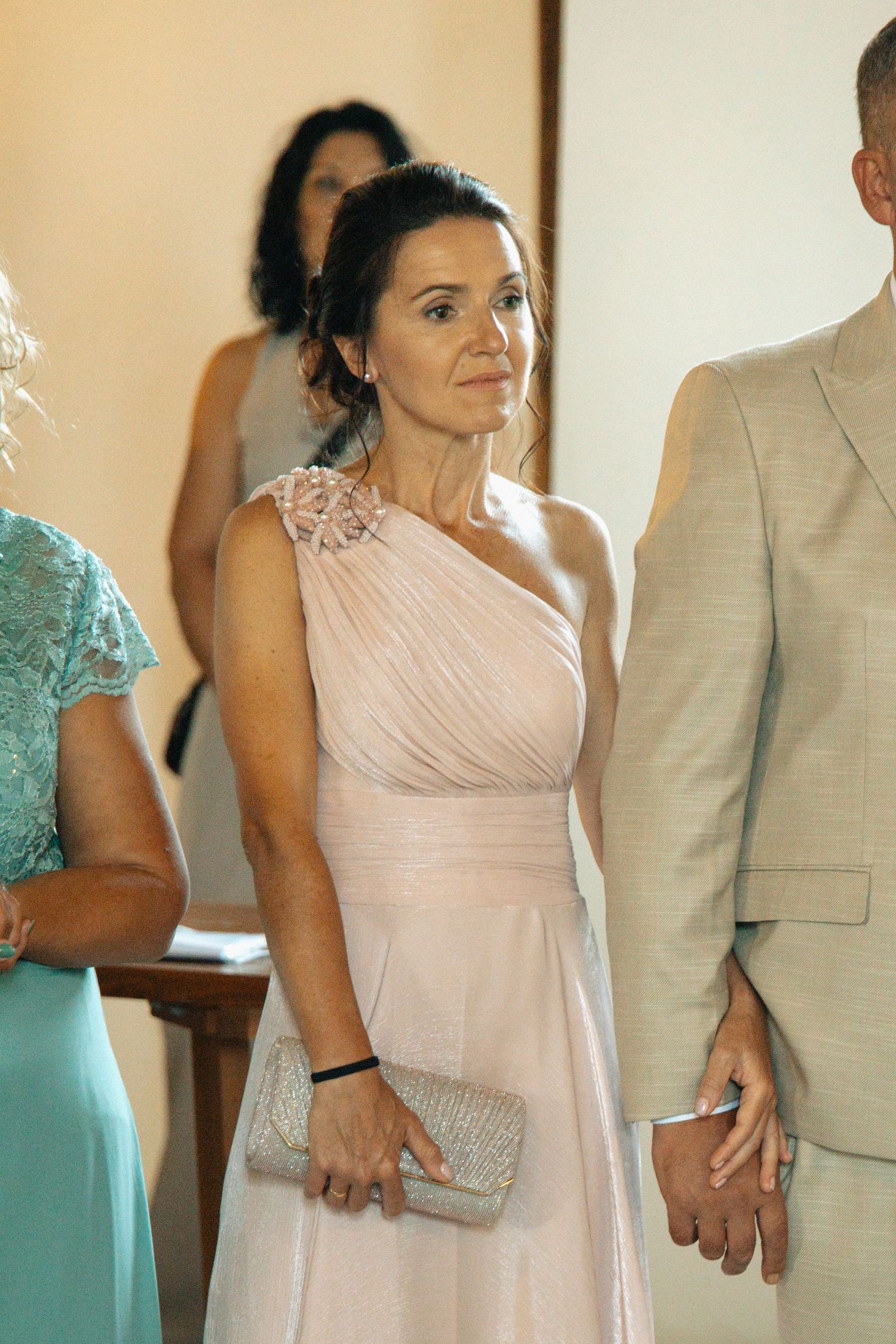 Elegant woman in blush one-shoulder gown holding clutch at a wedding.