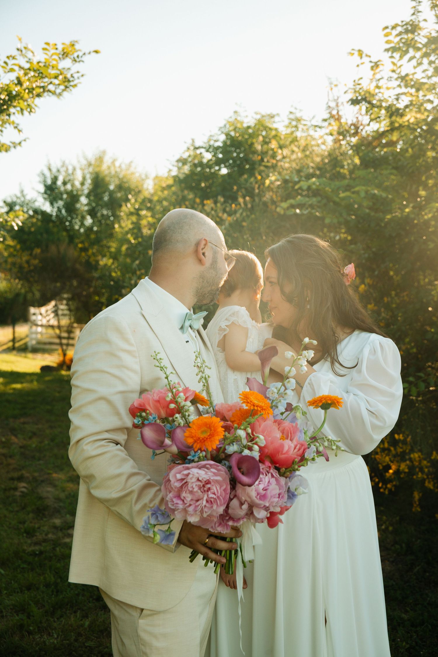 A couple in wedding attire holds a colorful bouquet while sharing a tender moment with their child outdoors.