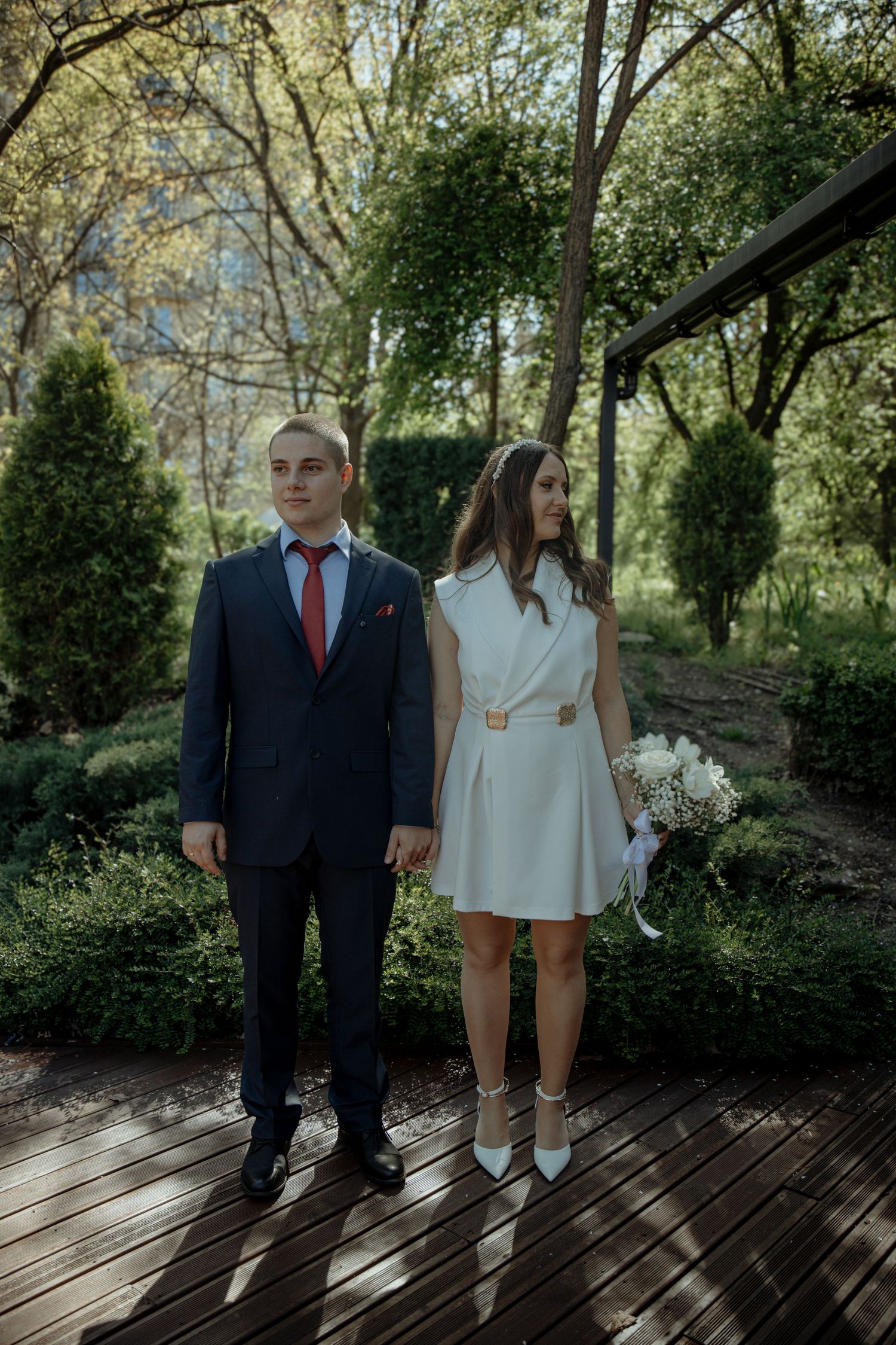 A couple in wedding attire stands on a wooden deck surrounded by lush greenery, bride holding a white bouquet.