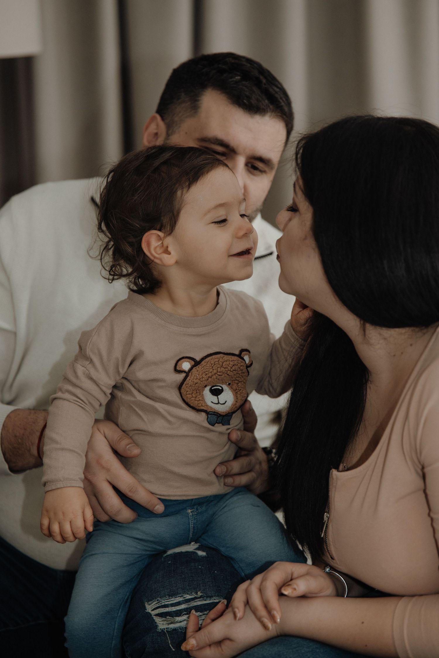 A young family sharing a tender moment, parents lovingly gazing at their toddler in a bear sweater indoors.