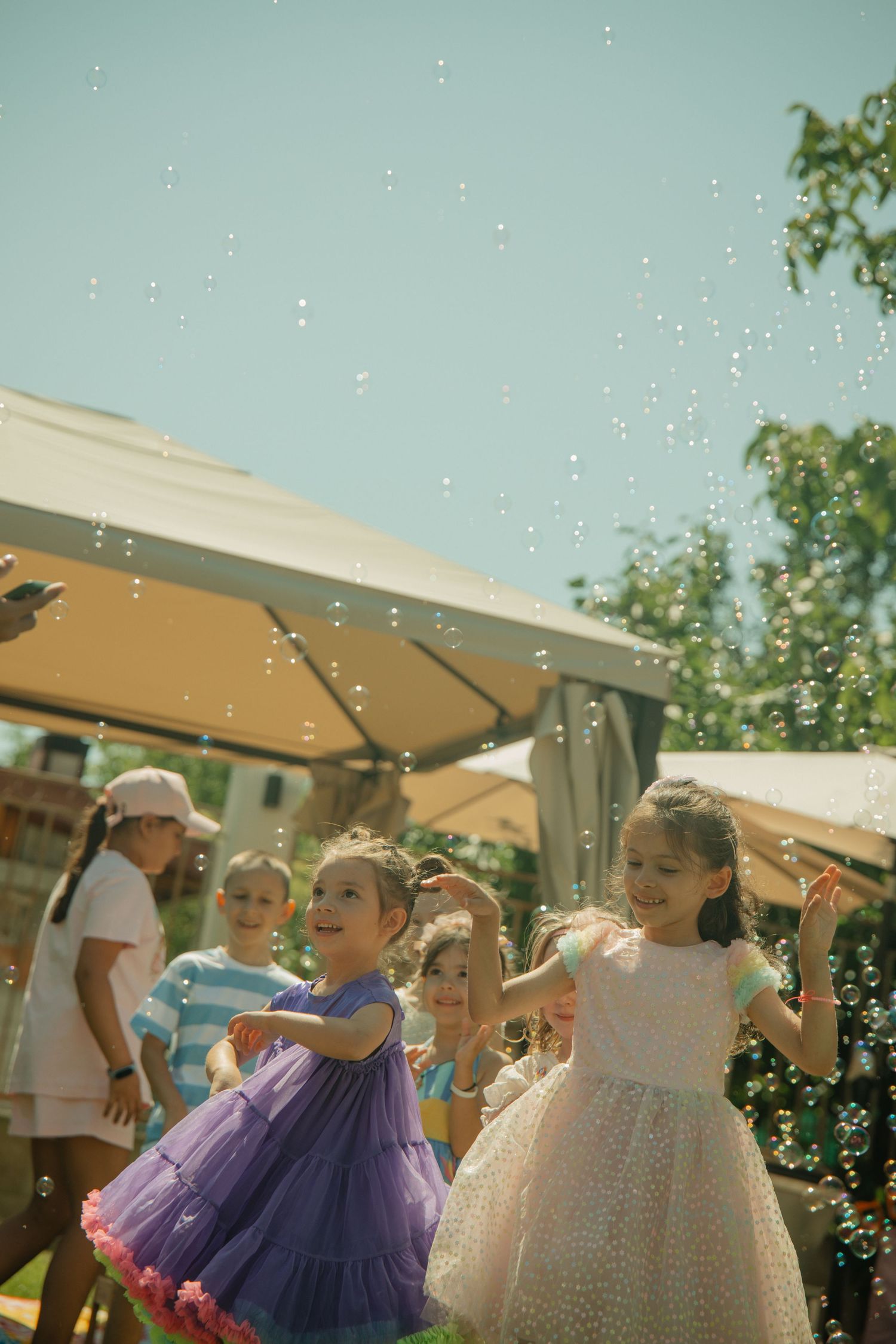 Children in colorful dresses playing with soap bubbles outdoors at a sunny garden party celebration.